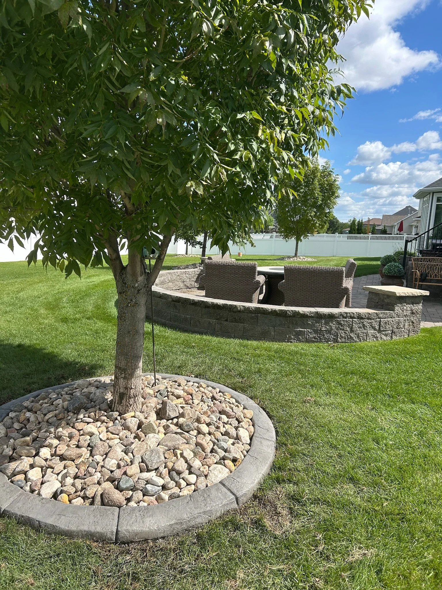 A backyard with a large green-leafed tree in a round stone-edged mulch bed, a brick patio with outdoor furniture, and a white fence under a blue sky with scattered clouds.