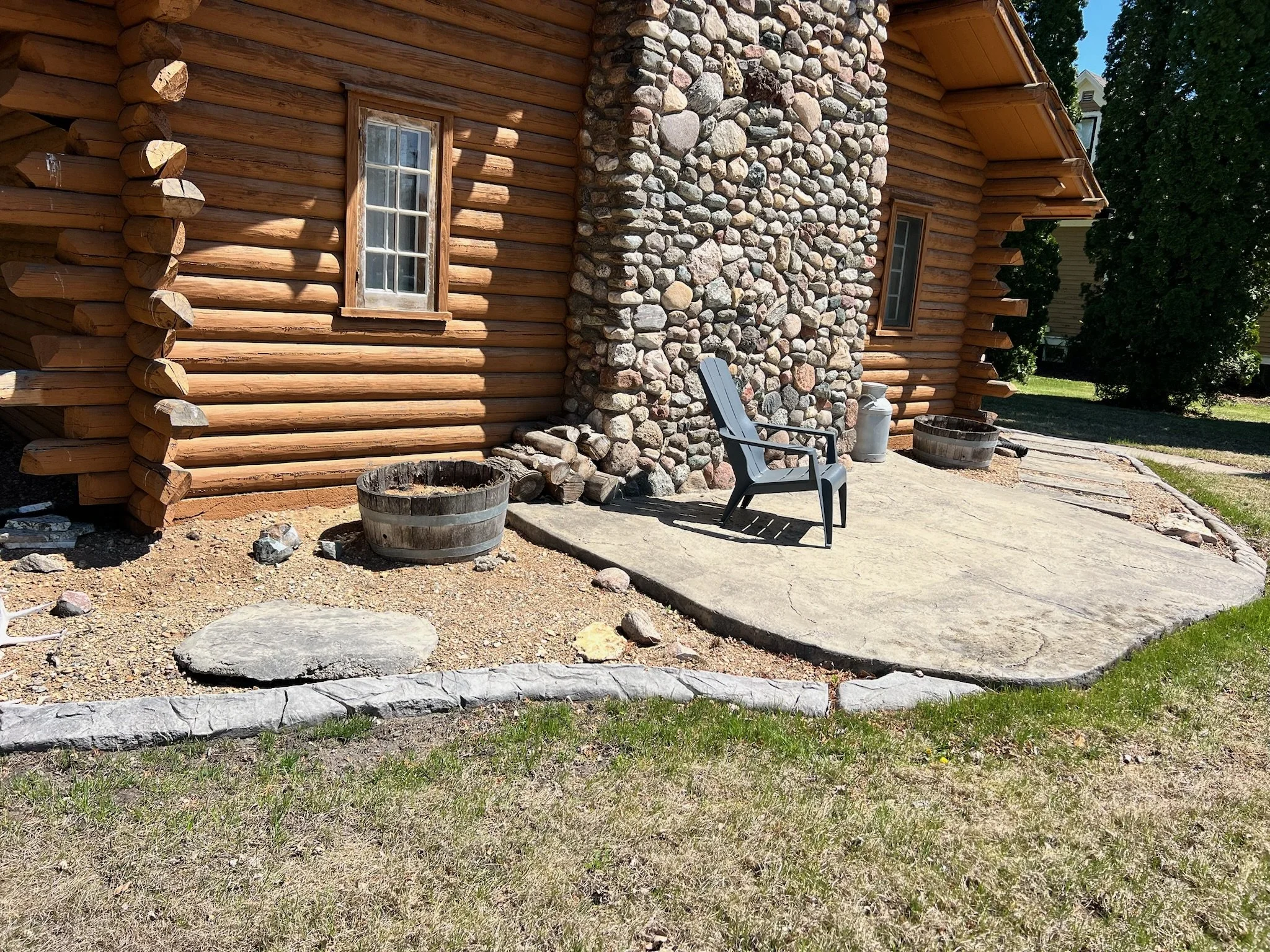 A small outdoor patio with a single gray plastic chair, surrounded by a rustic log cabin wall and a stone chimney, with some wooden planters and rocks, and a grassy yard.