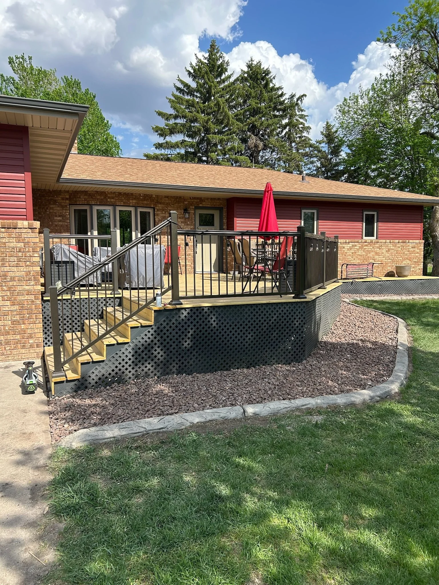 A backyard with a wooden deck attached to a red brick house, featuring outdoor furniture including a table with a red umbrella, a covered grill, and a few decorative planters, with a landscaped yard and large trees in the background.