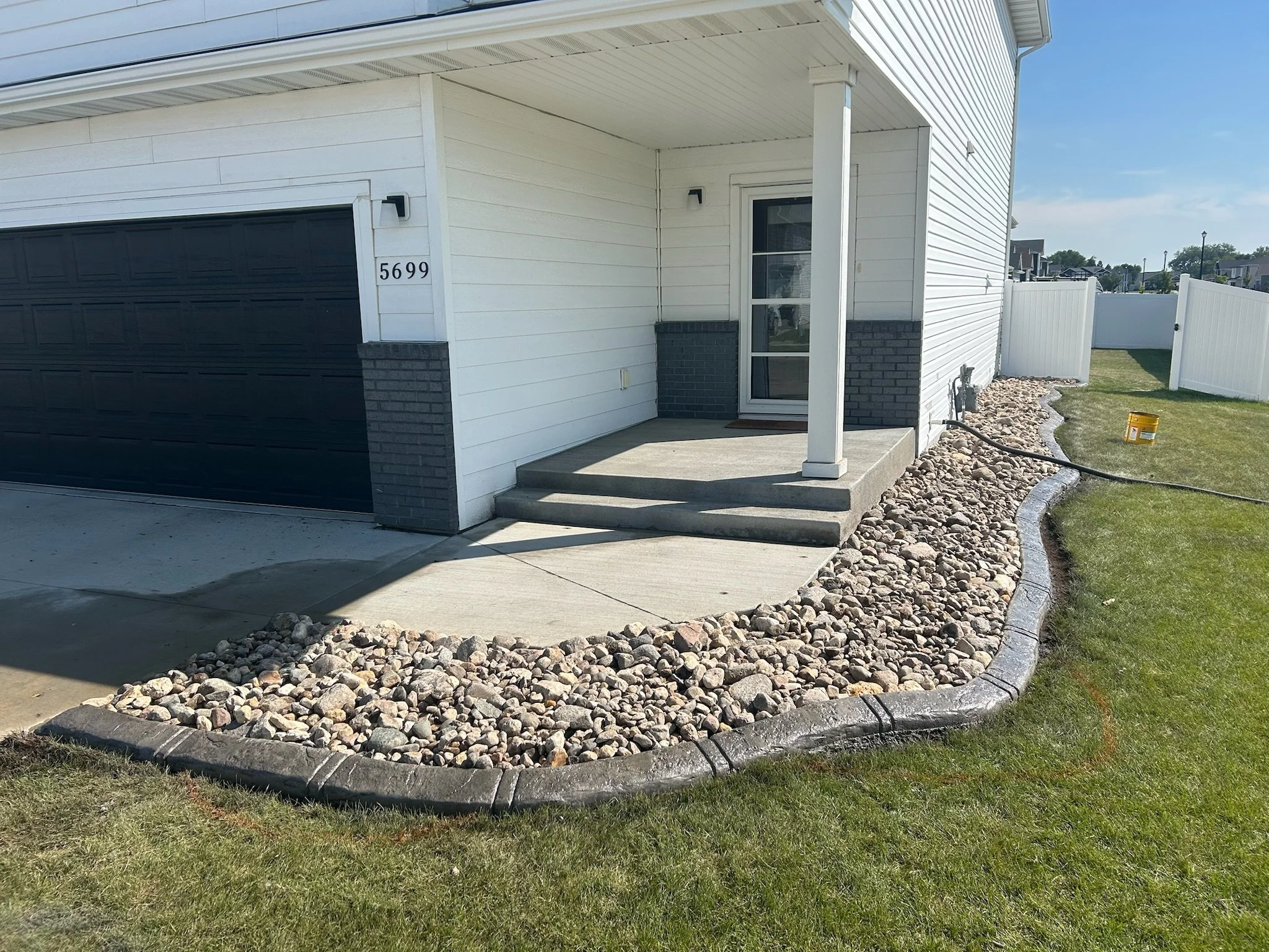 Front yard of a house with a small porch, concrete steps, and a landscaped area filled with rocks along the side of the house, with a lawn and white fence in the background.