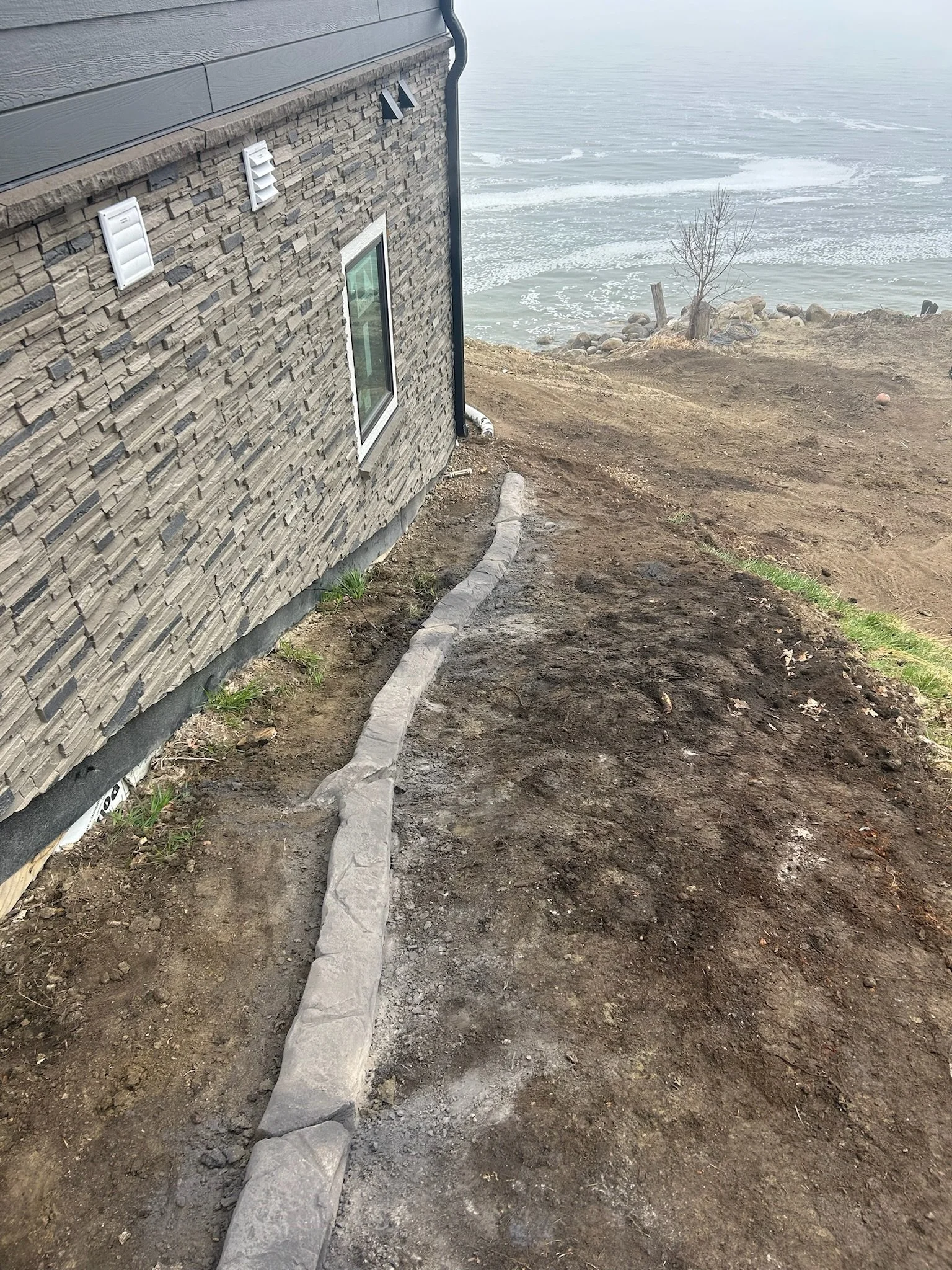 A house with gray siding and a stone exterior is next to a dirt path lined with stones, leading down to a rocky shoreline with water and small waves in the background.
