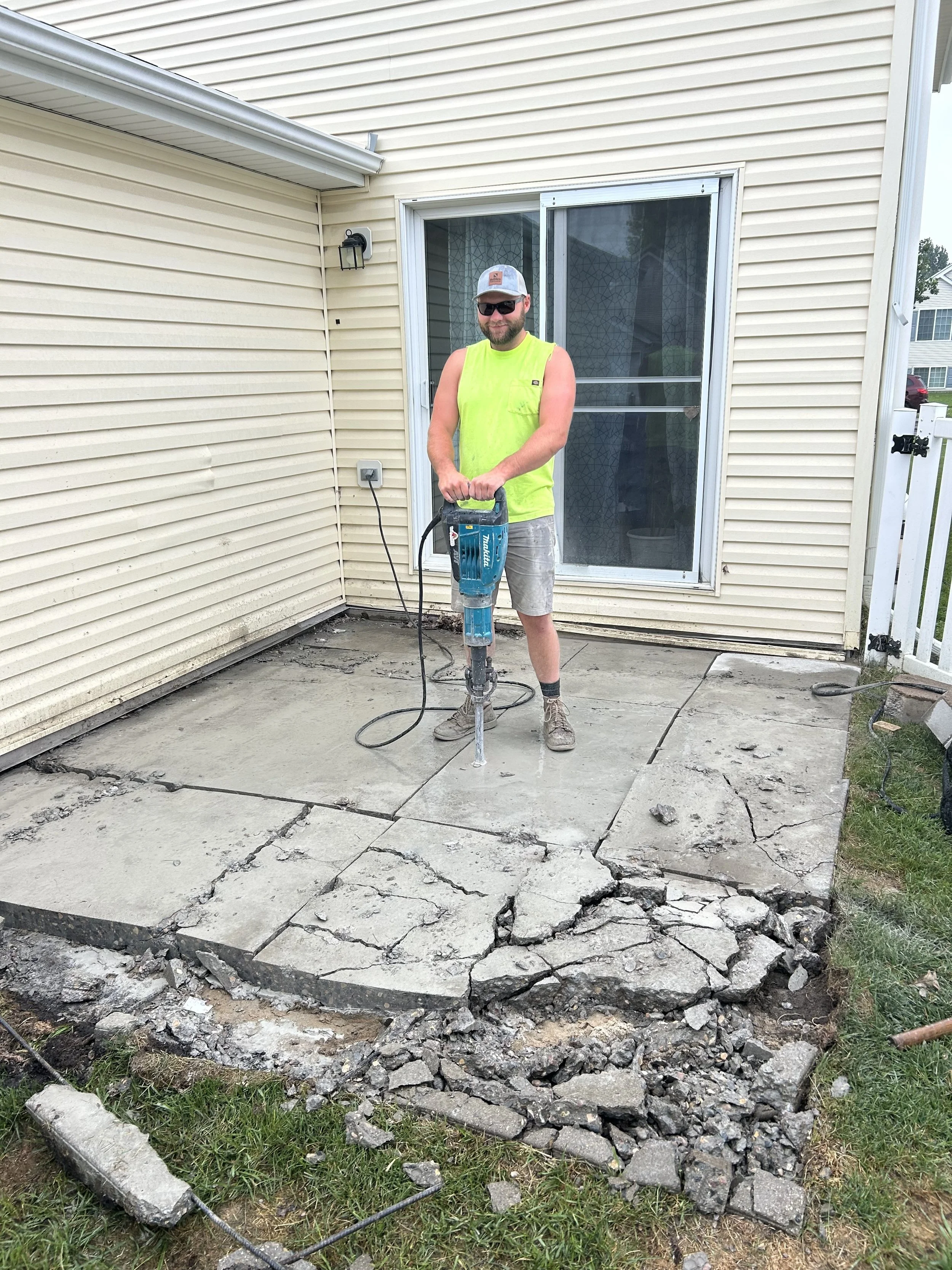 A man wearing sunglasses, a neon yellow sleeveless shirt, and gray shorts is using a jackhammer to break up concrete on a patio outside a house. The house has beige siding, a sliding glass door, and a small fence.