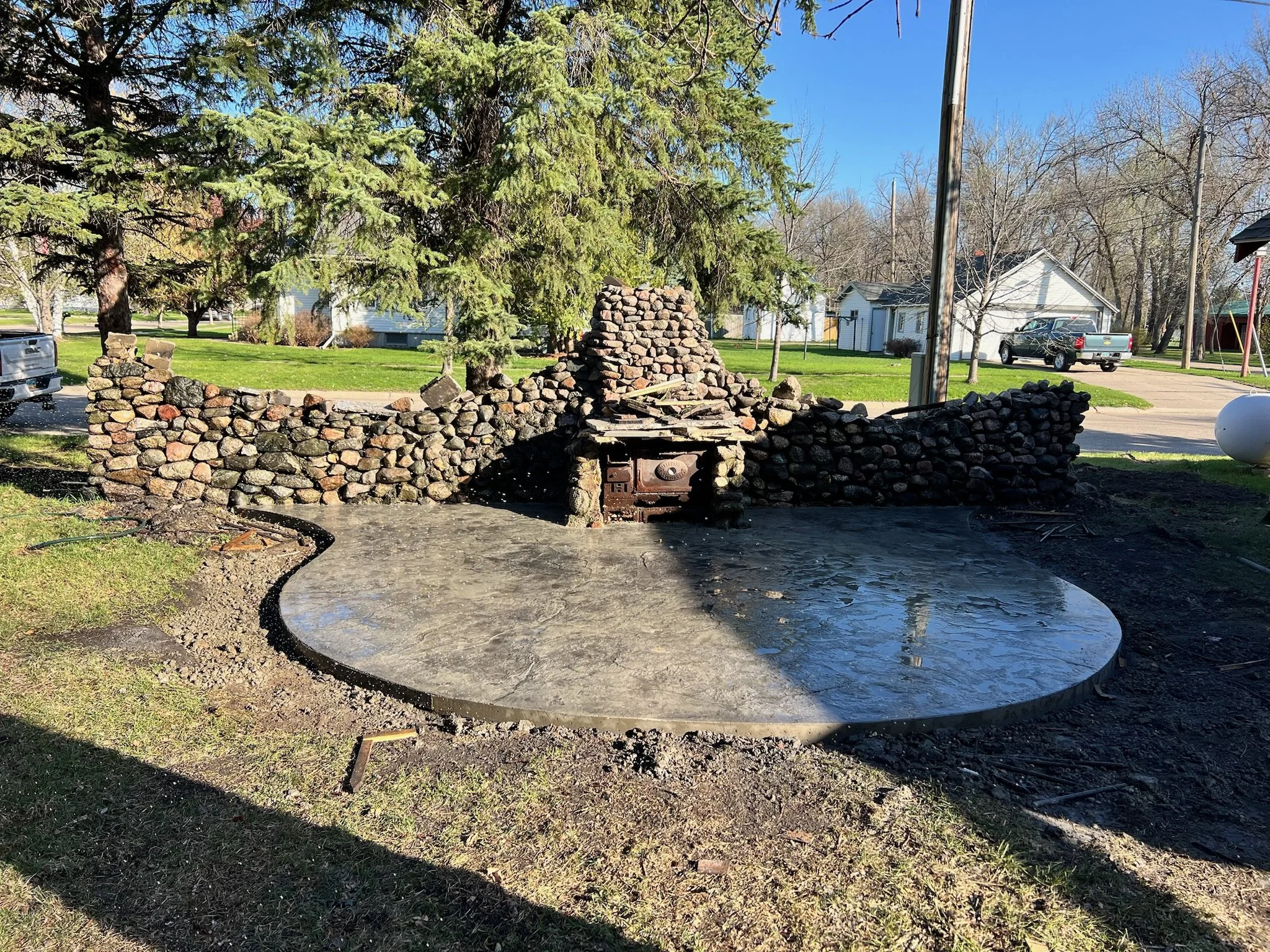 Stone fire pit with a conical chimney and a metal door, surrounded by a partially paved circular area, in a backyard with trees and houses in the background.