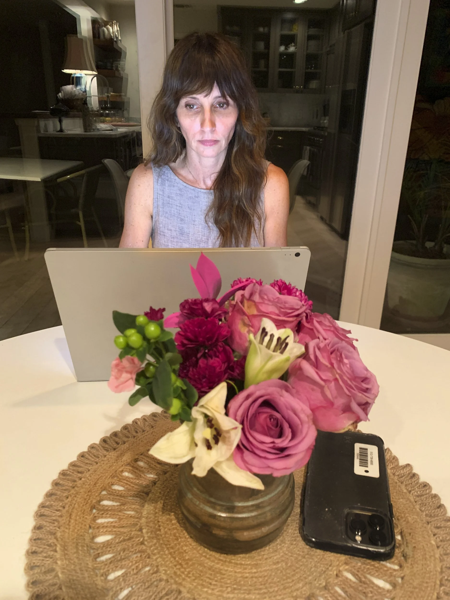 Smith Bohls, "Motherhood," 2022. A mom is working on replying to emails and finishing up work in her home environment. She is sitting at her kitchen table in a familiar space working.