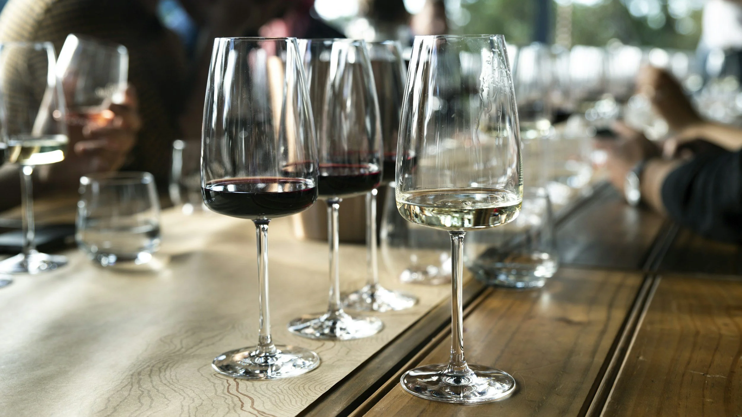A row of wine glasses on a wooden table, with some filled with red wine, some with white wine, and others empty, in a social setting.