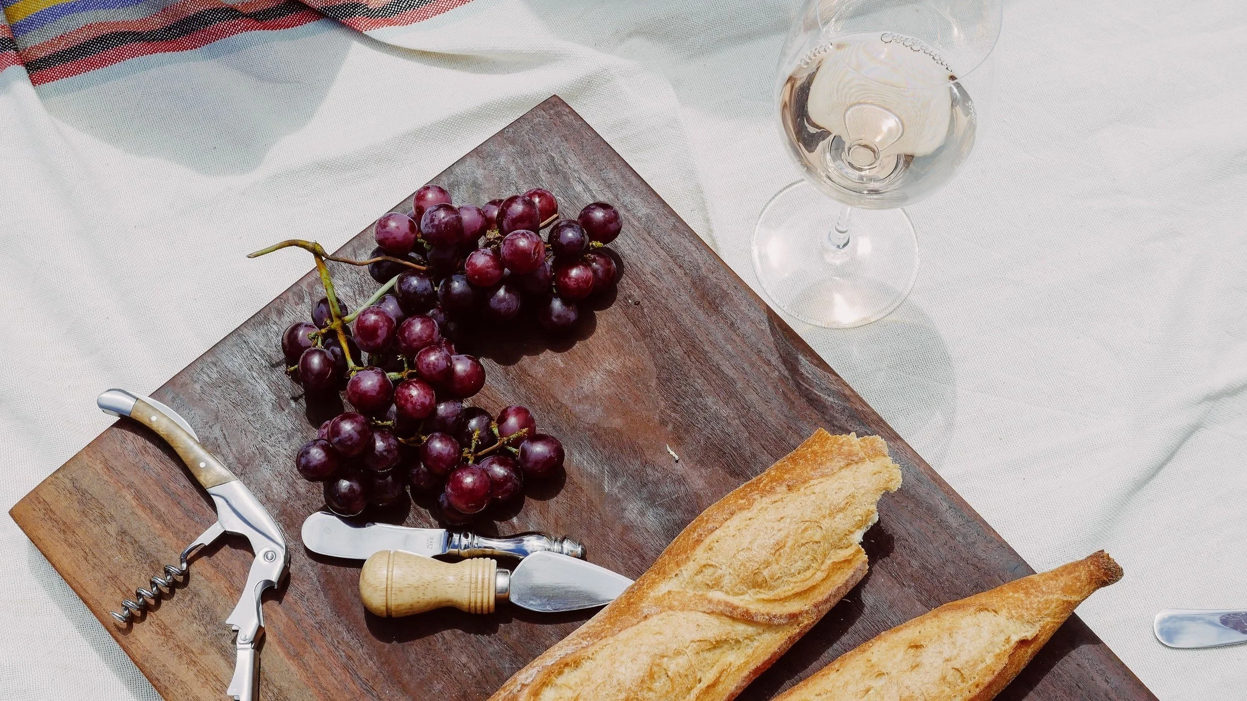 A bunch of red grapes on a wooden cutting board, a cracked baguette, a corkscrew, a small cheese knife, and a glass of white wine on a white tablecloth.