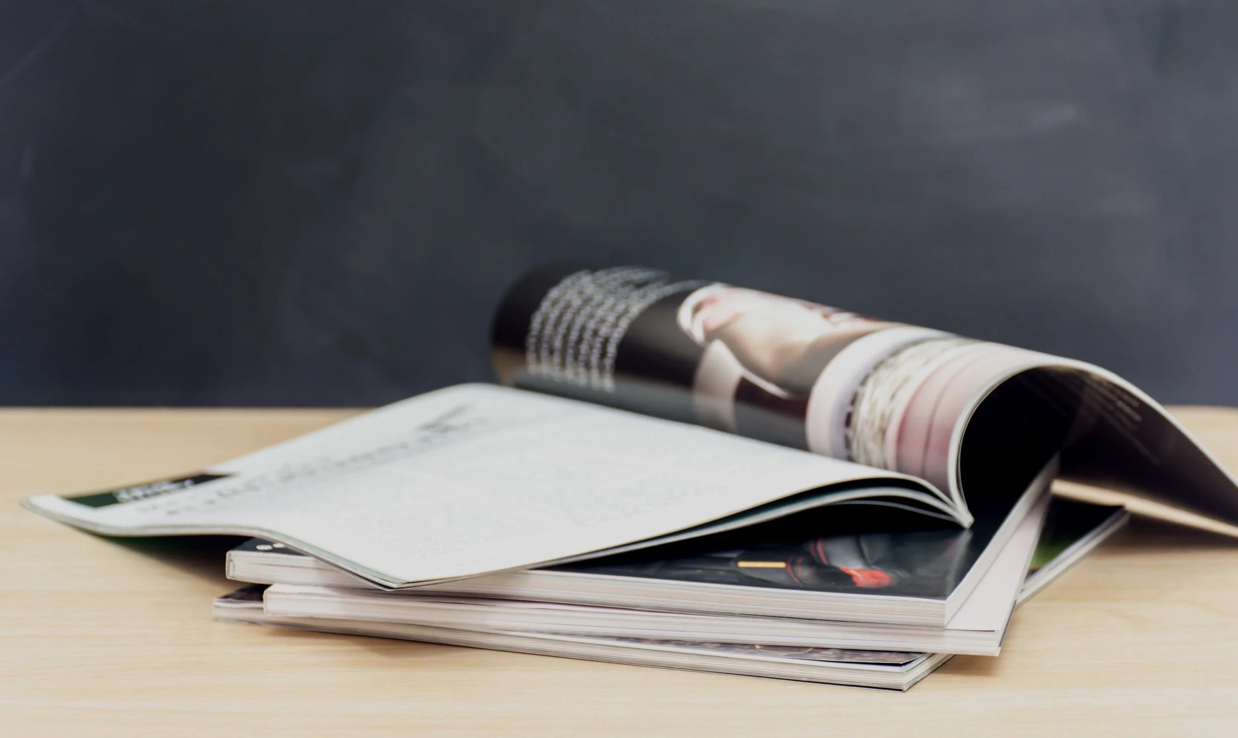 A stack of magazines and newspapers on a wooden table, with one magazine open and some pages slightly curled.