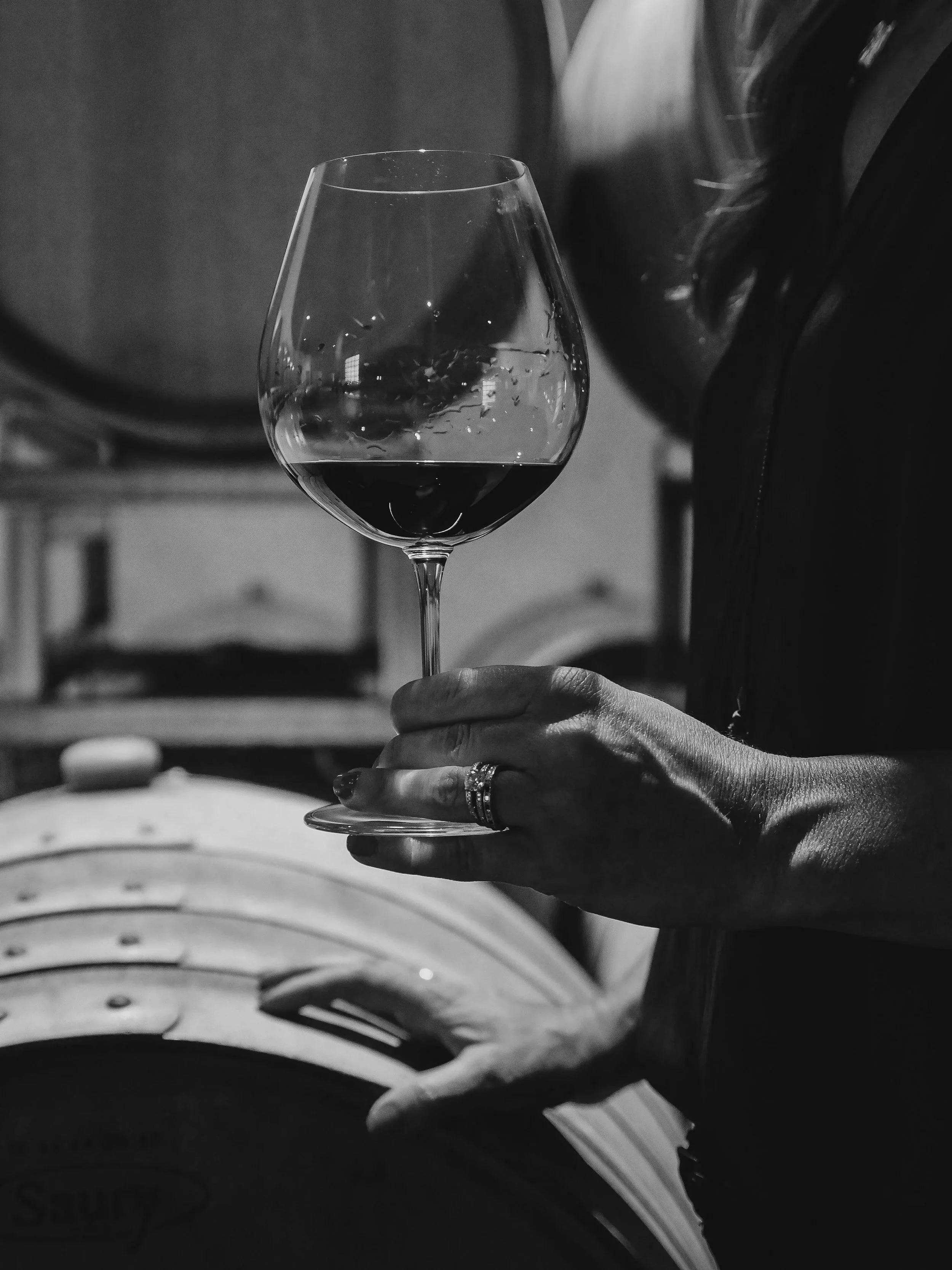 Person holding a glass of red wine in a winery or cellar with wine barrels in the background, black and white photo.