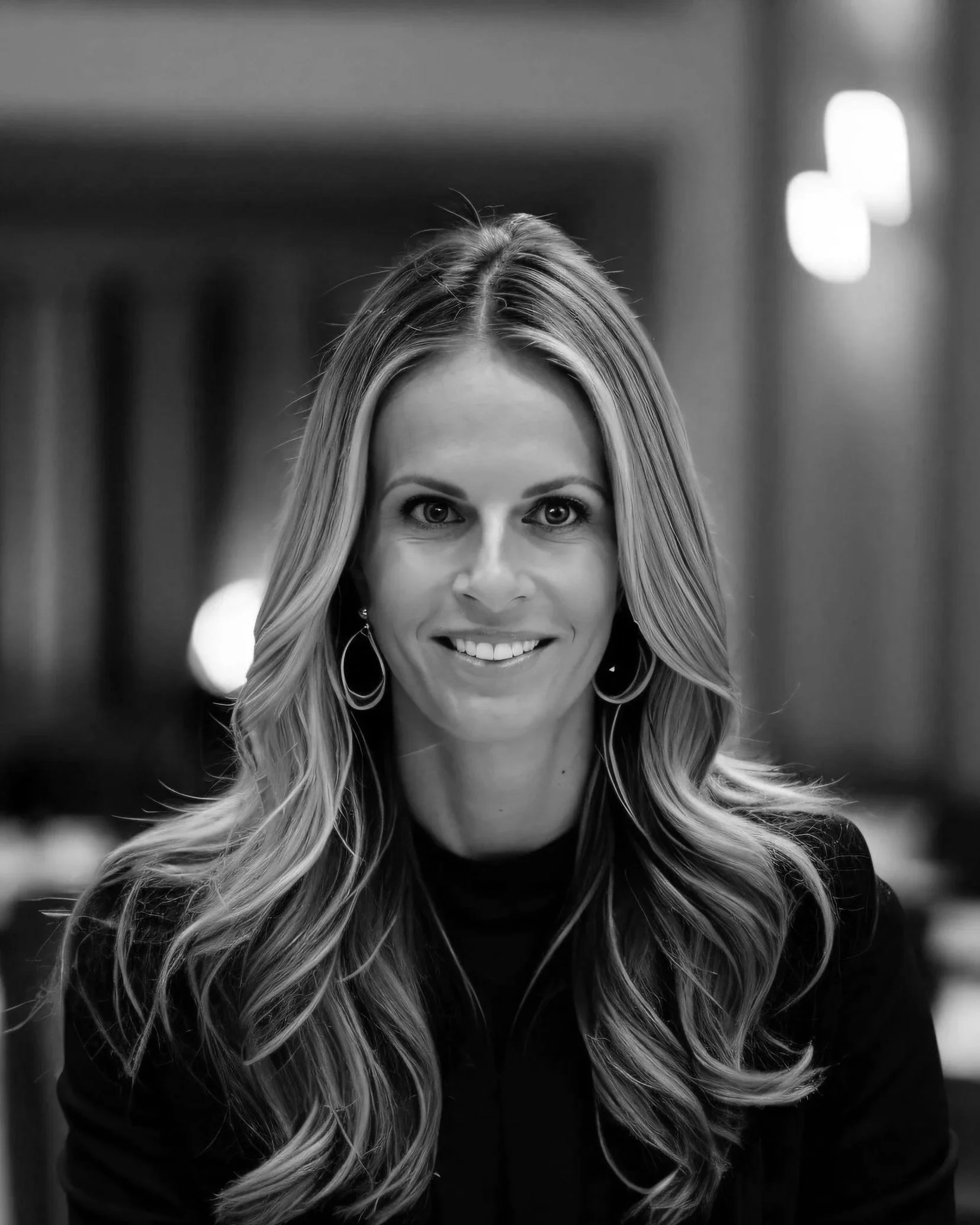 Black and white photo of a woman with long wavy hair, smiling, wearing earrings.