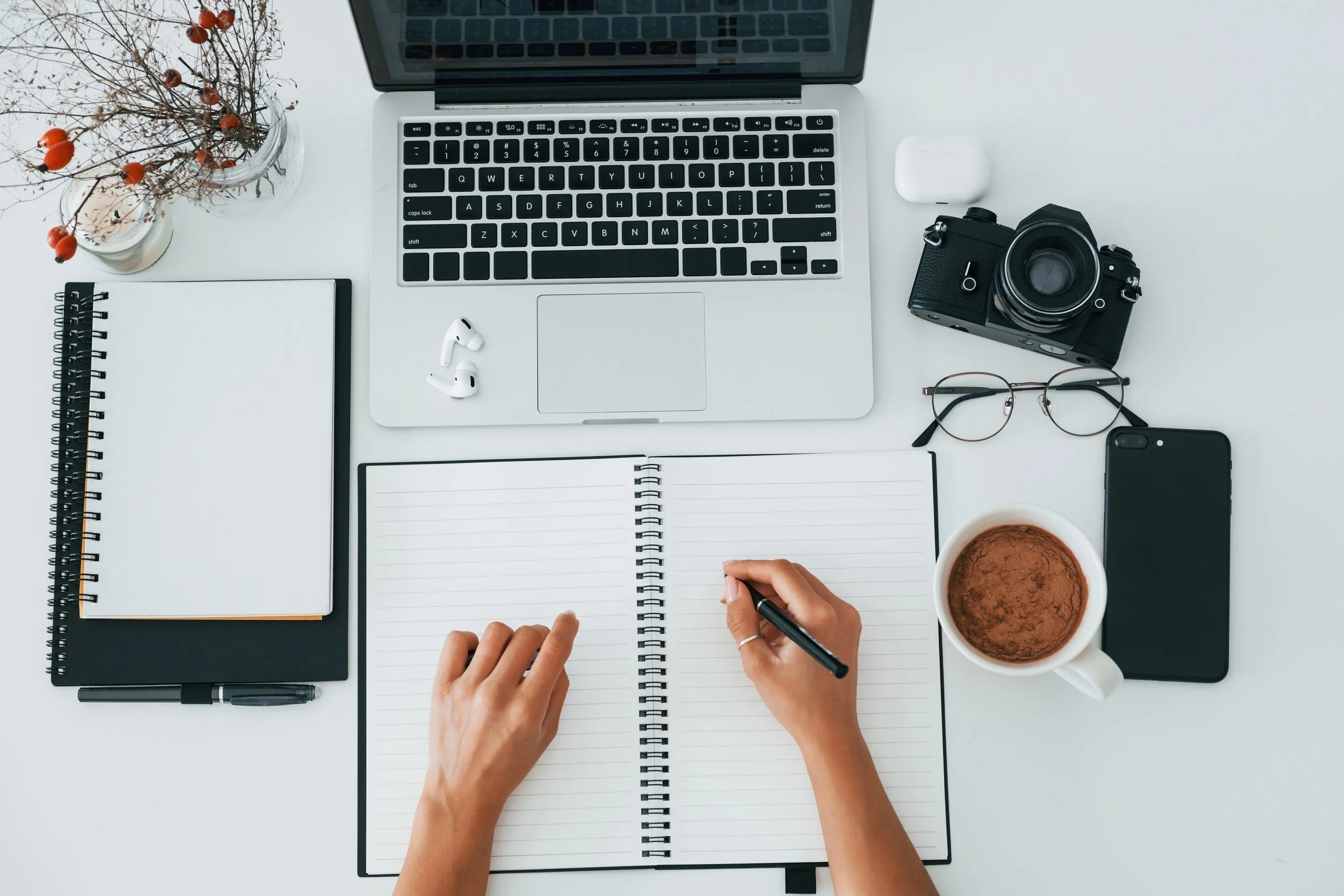 Top view of a white desk with a person writing in a lined notebook, surrounded by a laptop, camera, glasses, smartphone, coffee mug, earphones, a small notebook, a pen, and a vase of dried flowers.