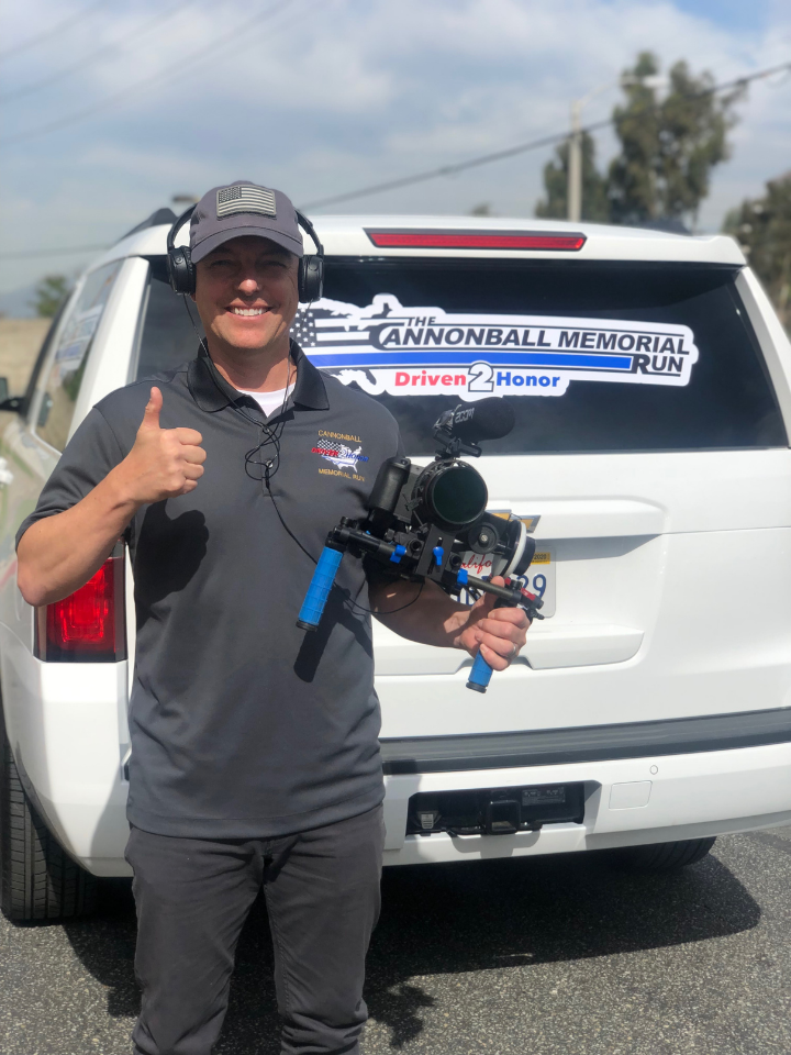 Christopher Lee filming the Cannonball Memorial Run, standing beside a vehicle with the event logo, documenting the cross-country tribute in The Story Comes First.