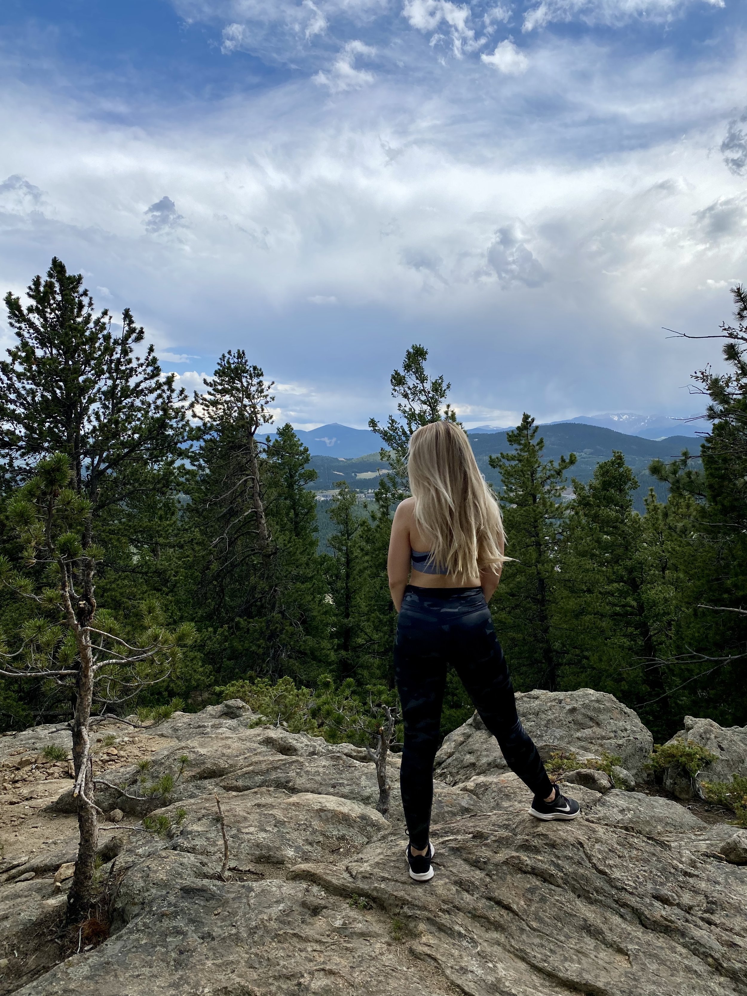 Woman with long blonde hair standing on a rocky hilltop, surrounded by trees and overlooking a mountain landscape under a cloudy sky.