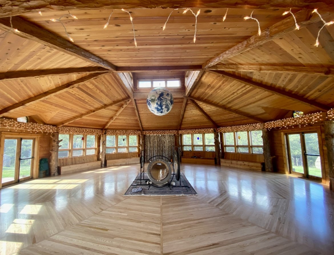 Spacious wooden octagonal room with vaulted ceiling, central hanging globe, large gong, hardwood floors, string lights, and numerous windows.