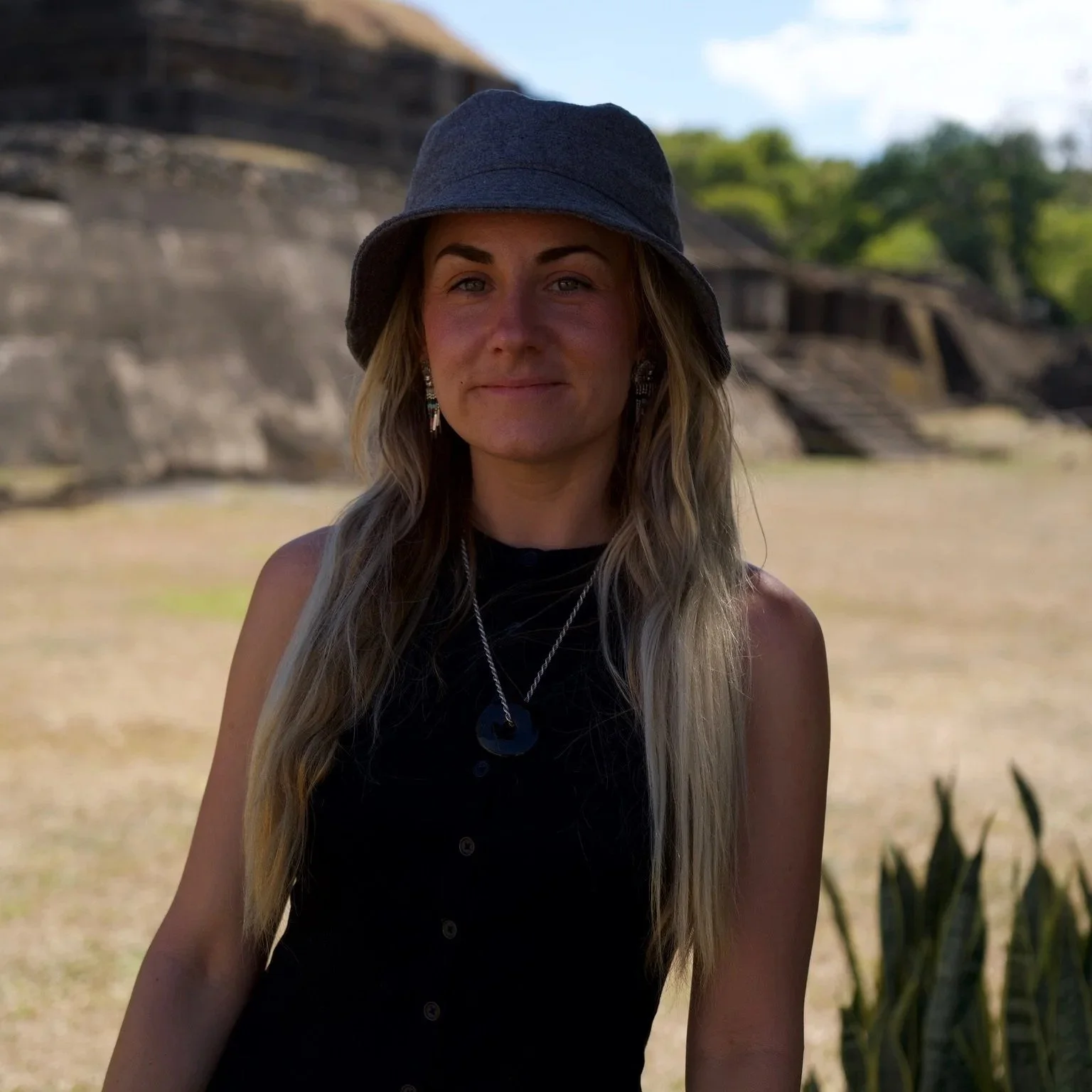 A woman with long blonde hair wearing a dark gray hat, black sleeveless top, and silver jewelry, standing outdoors with a rocky cliff and blue sky in the background.