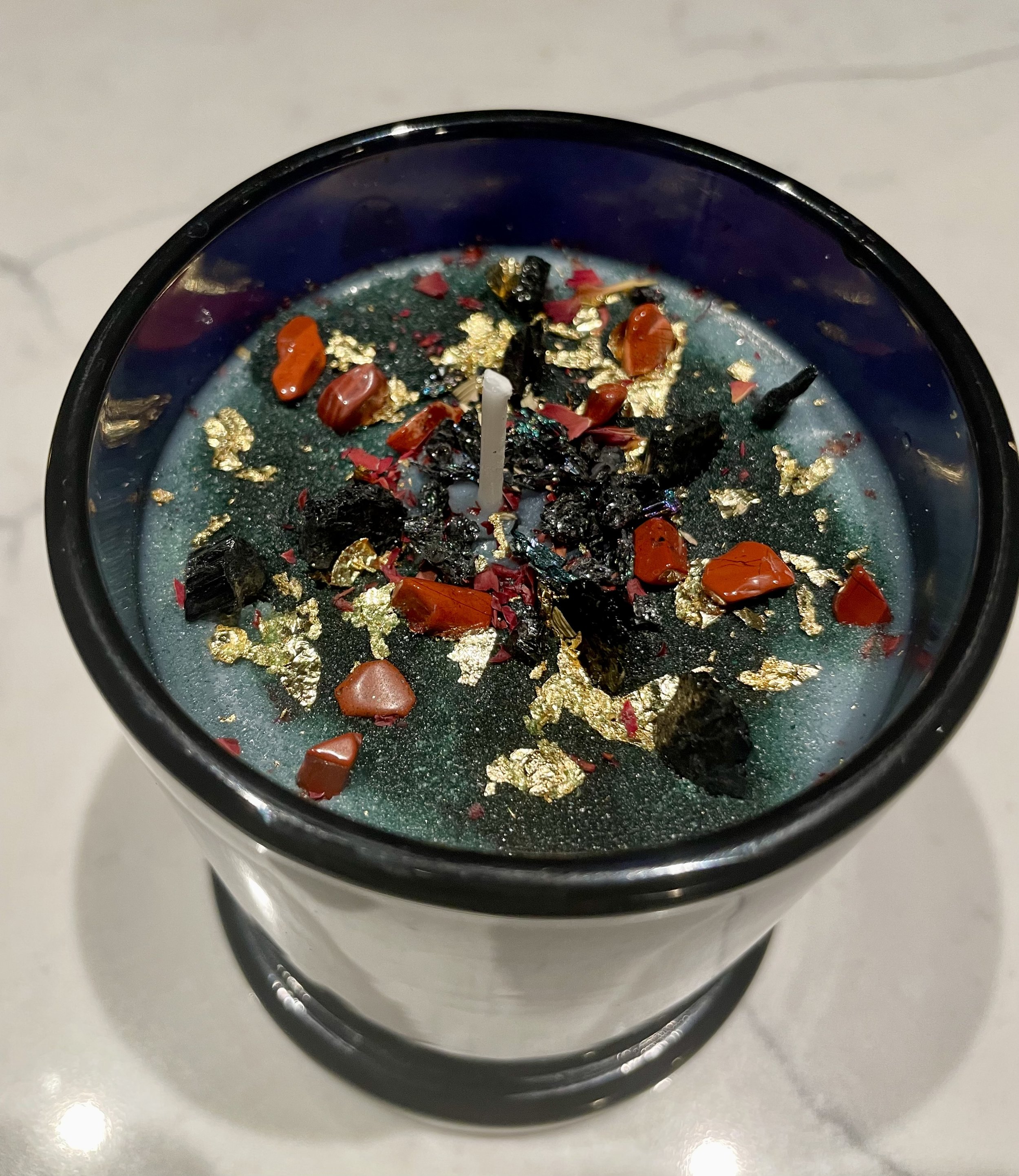 Decorative candle with gemstones, gold flakes, and dark wax in a black container.