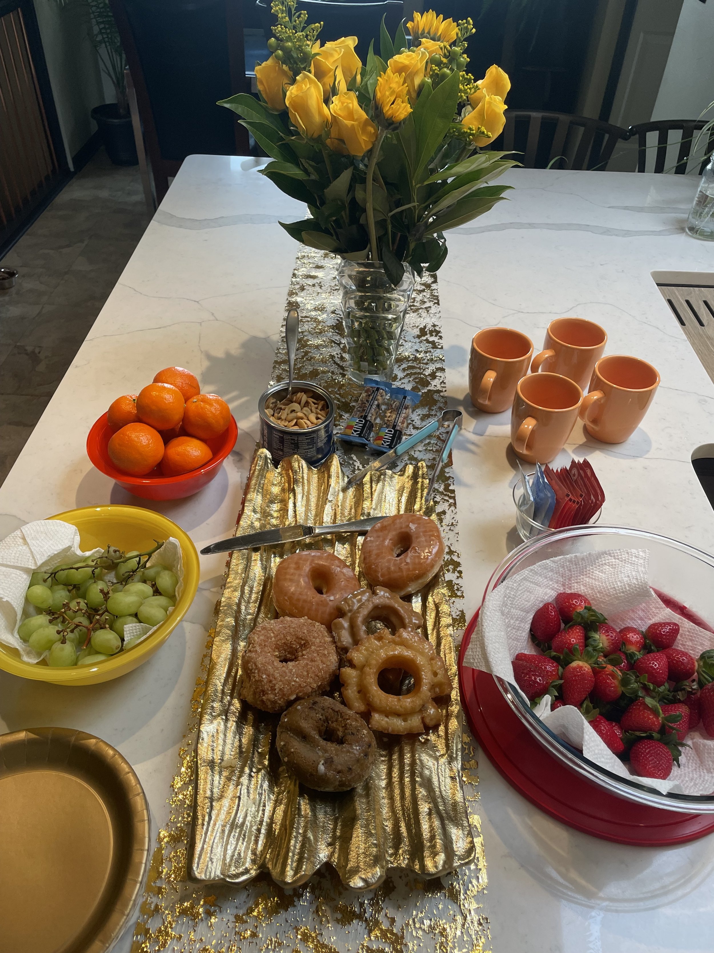 Kitchen counter with a display of assorted donuts on a gold tray, bowls of strawberries, grapes, and oranges, mixed nuts, a vase of yellow flowers, and several orange mugs.