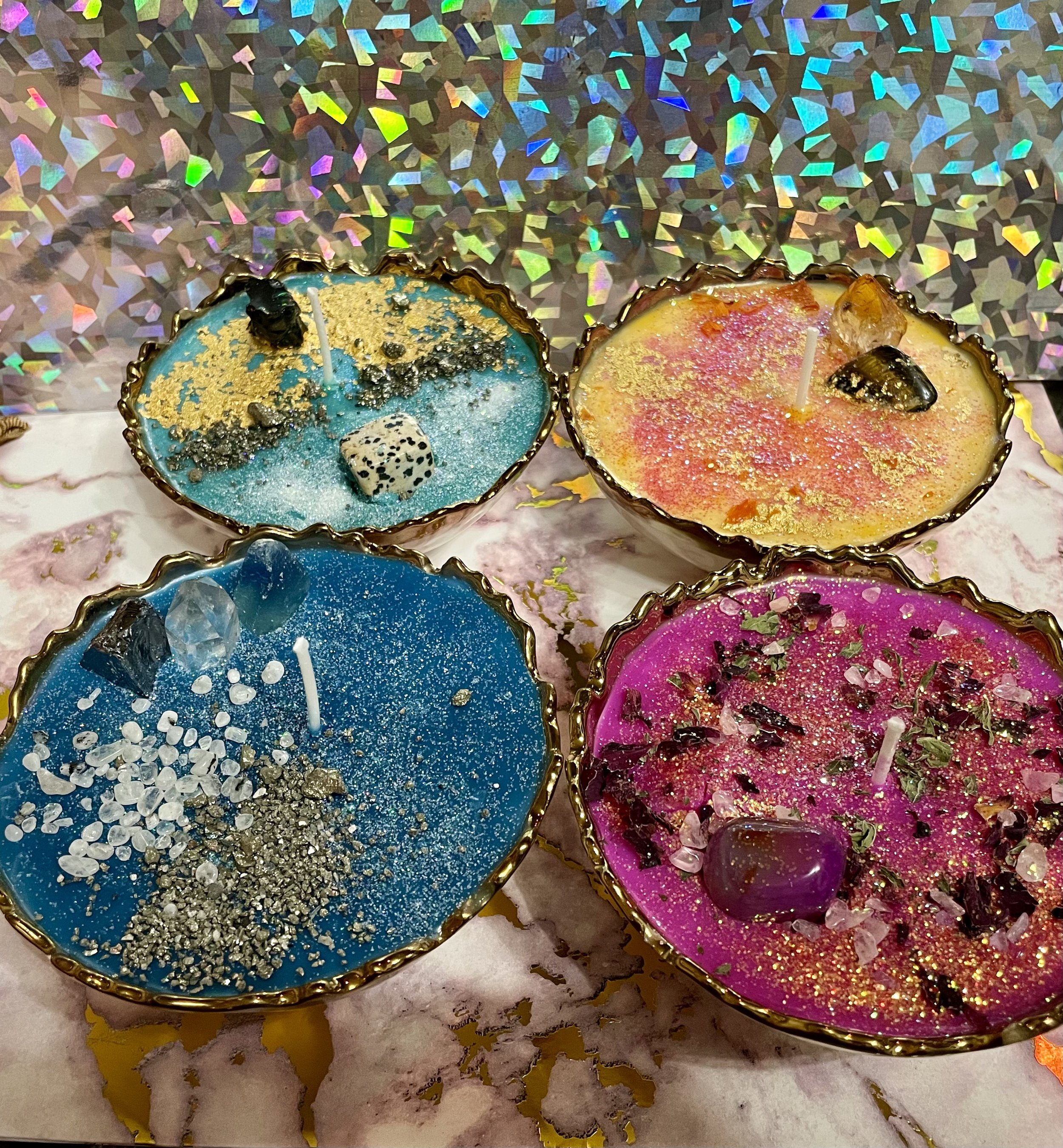 Four colorful decorative candles with gemstones and glitter, displayed on a marble surface with holographic background.