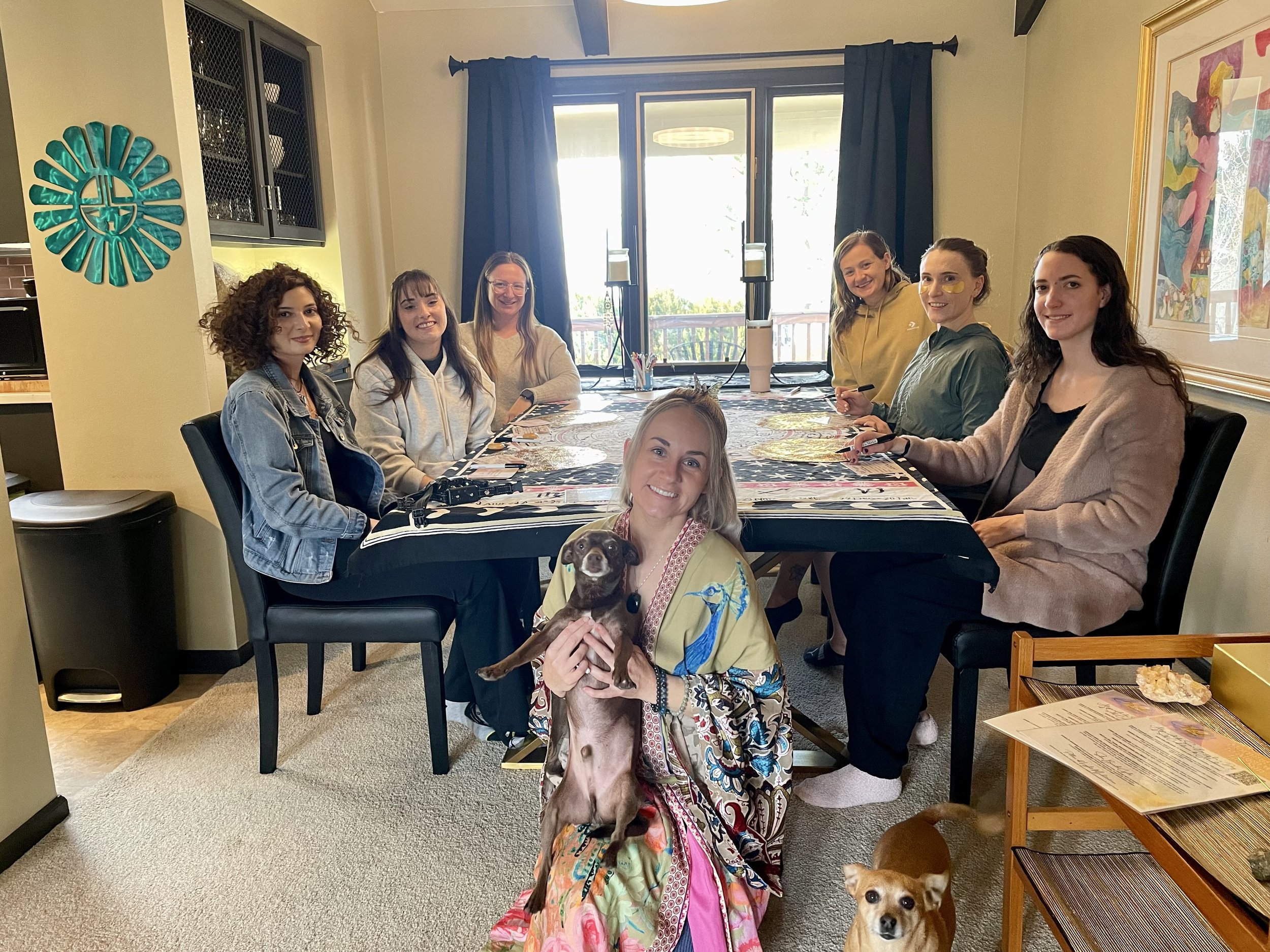 A group of seven women sitting around a dining table, engaged in an activity, with a dog sitting on the lap of one woman in the foreground. Another small dog is on the floor. The room has large windows, a decorative wall art piece, and a framed color