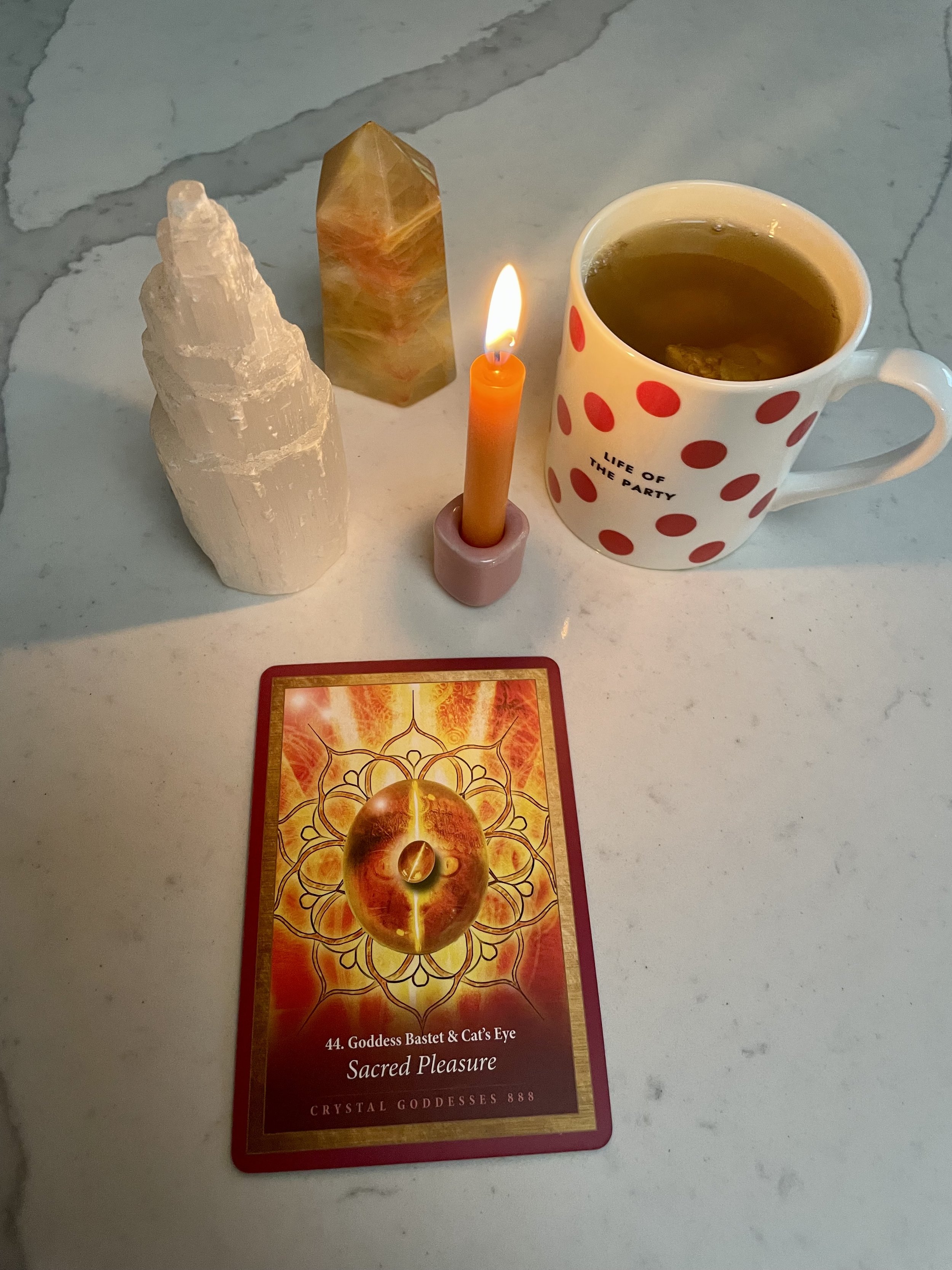 Scene with a white selenite crystal tower, amber crystal point, glowing candle, polka dot mug with tea, and an oracle card titled 'Sacred Pleasure' featuring Goddess Bastet & Cat’s Eye design on a table.