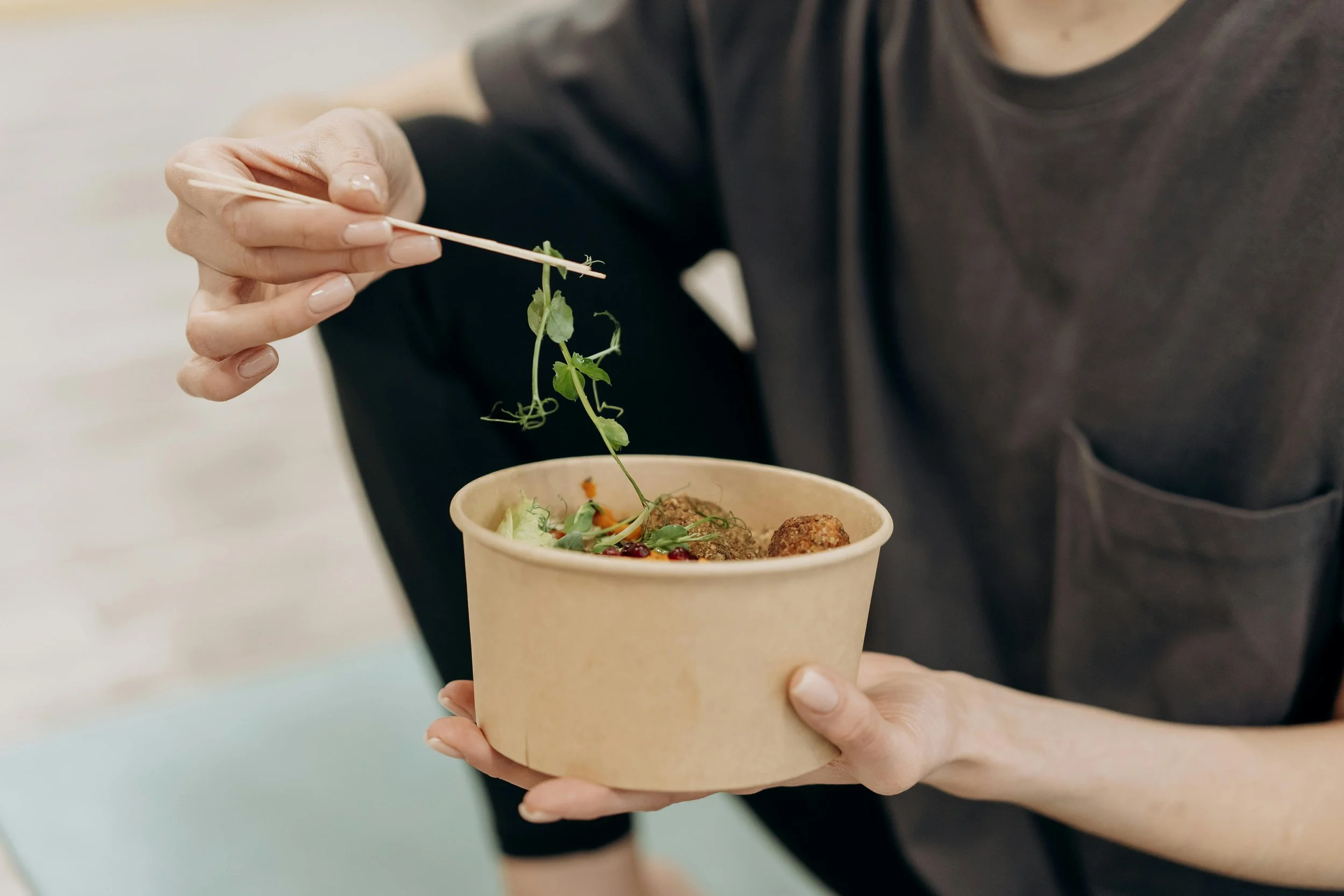 Person holding a paper bowl with salad and using chopsticks to pick herbs.
