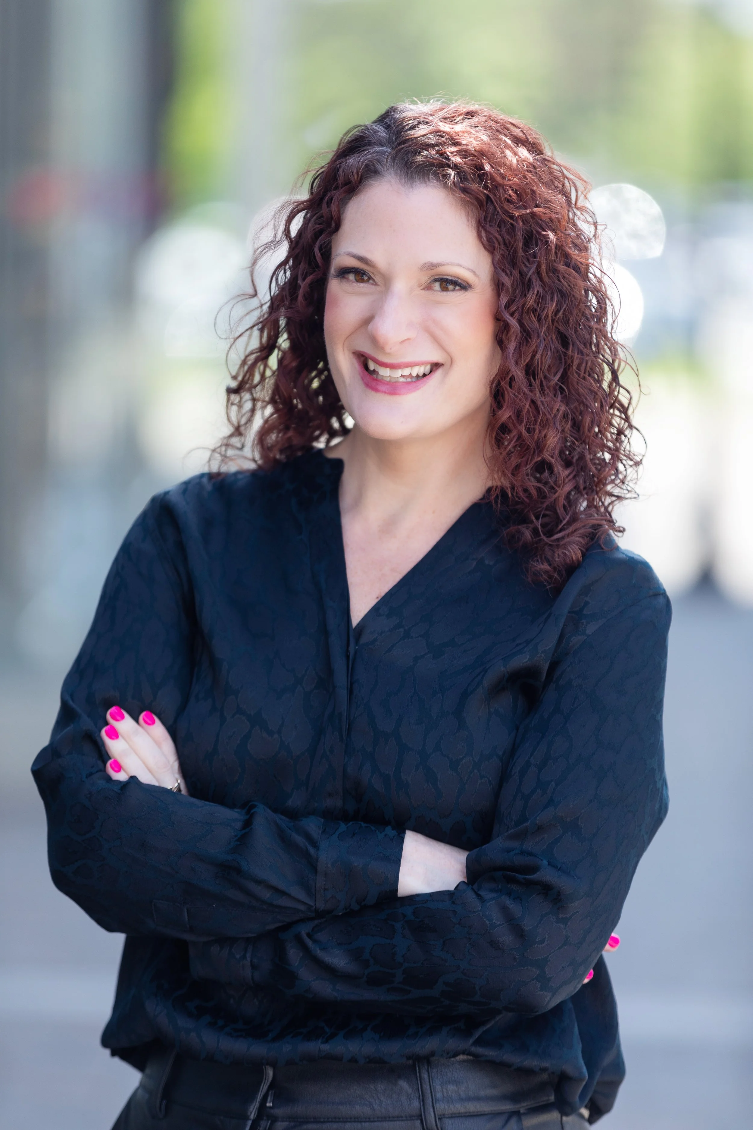 A woman with curly red hair and pink lipstick, standing outdoors with her arms crossed, smiling at the camera, wearing a dark blue patterned blouse.