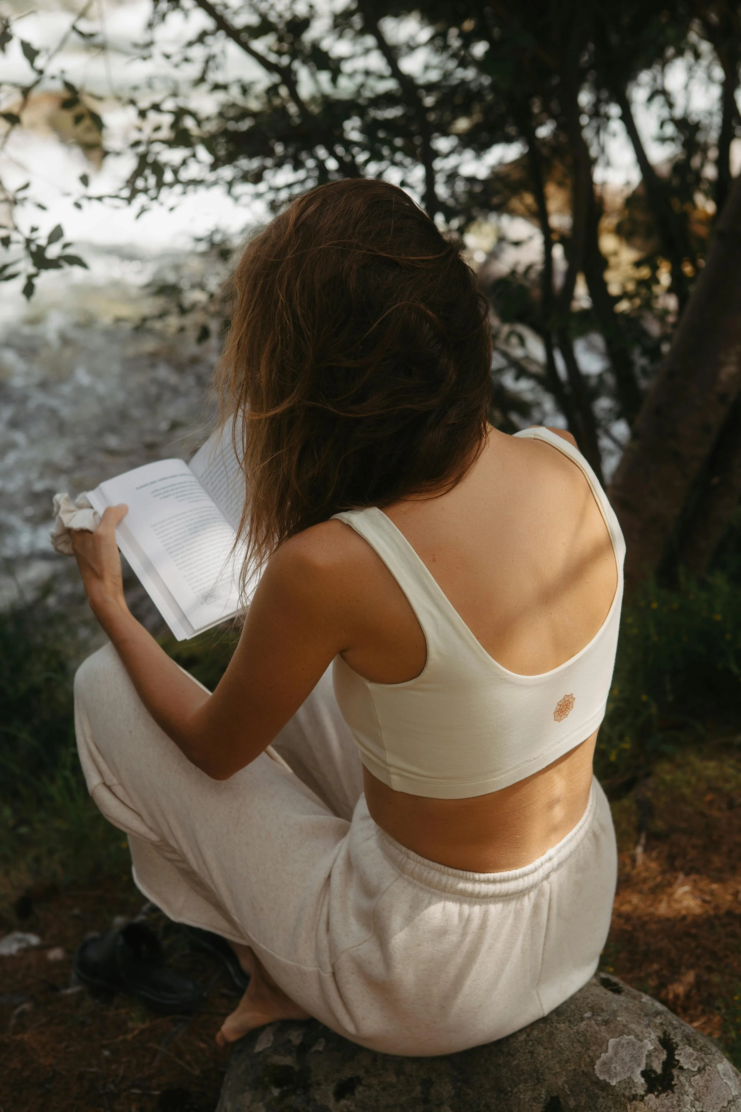 A person with brown hair, wearing a white sleeveless top and beige pants, sitting on a rock outdoors, reading a book with trees and water in the background.