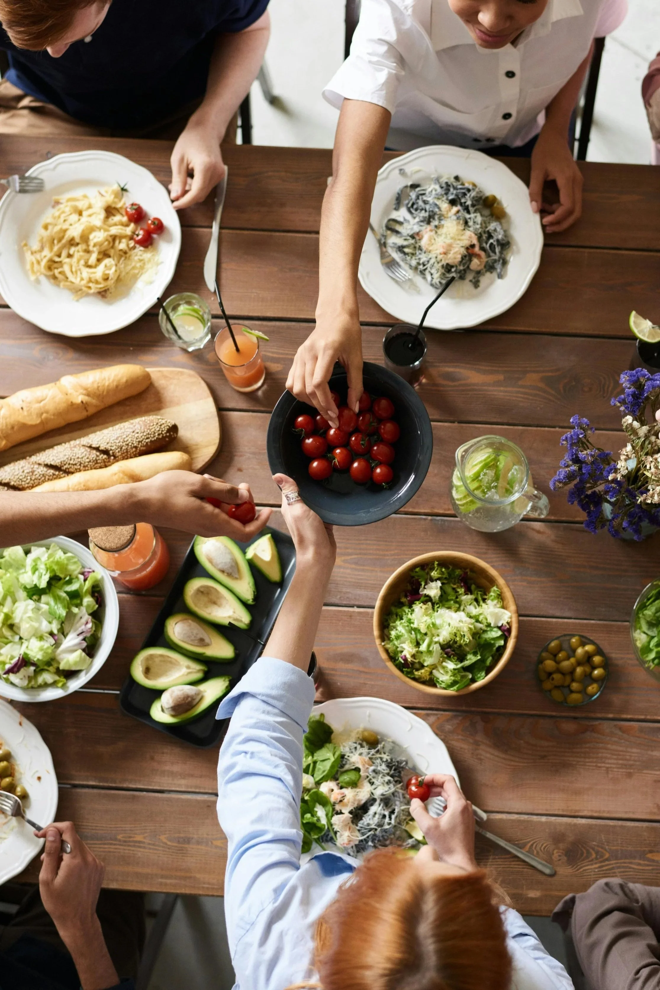 People sharing a meal of pasta, salad, bread, and beverages at a wooden dining table.