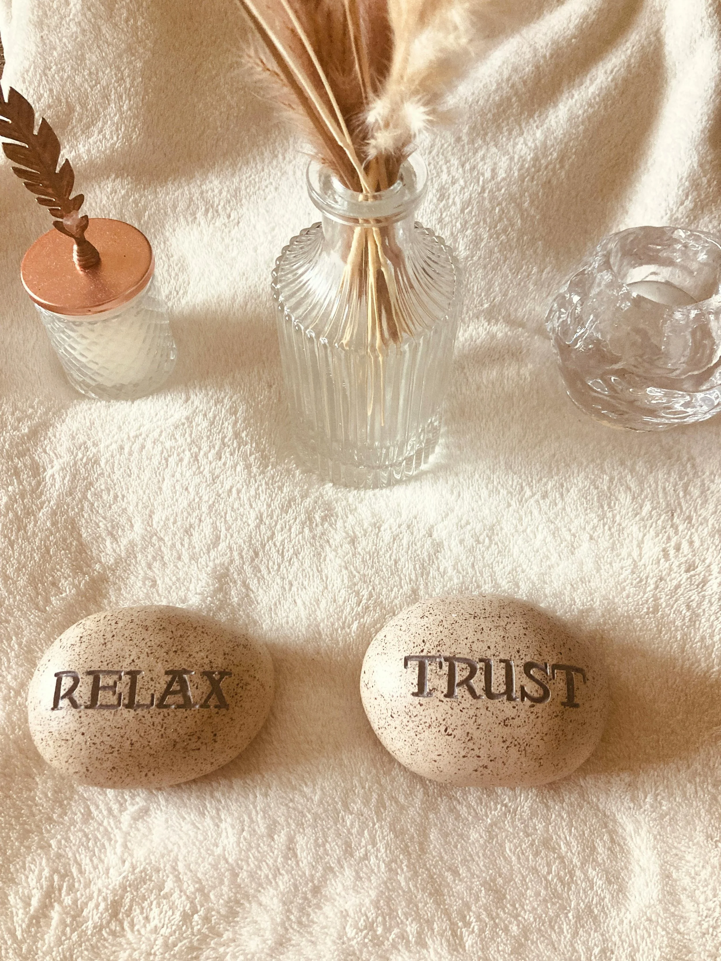 Two speckled stones with the words 'RELAX' and 'TRUST' written on them, placed on a soft beige surface. Behind them are three glass vases, one with a copper lid and a feather, another with dried pampas grass, and the third with a carved transparent glass sculpture.