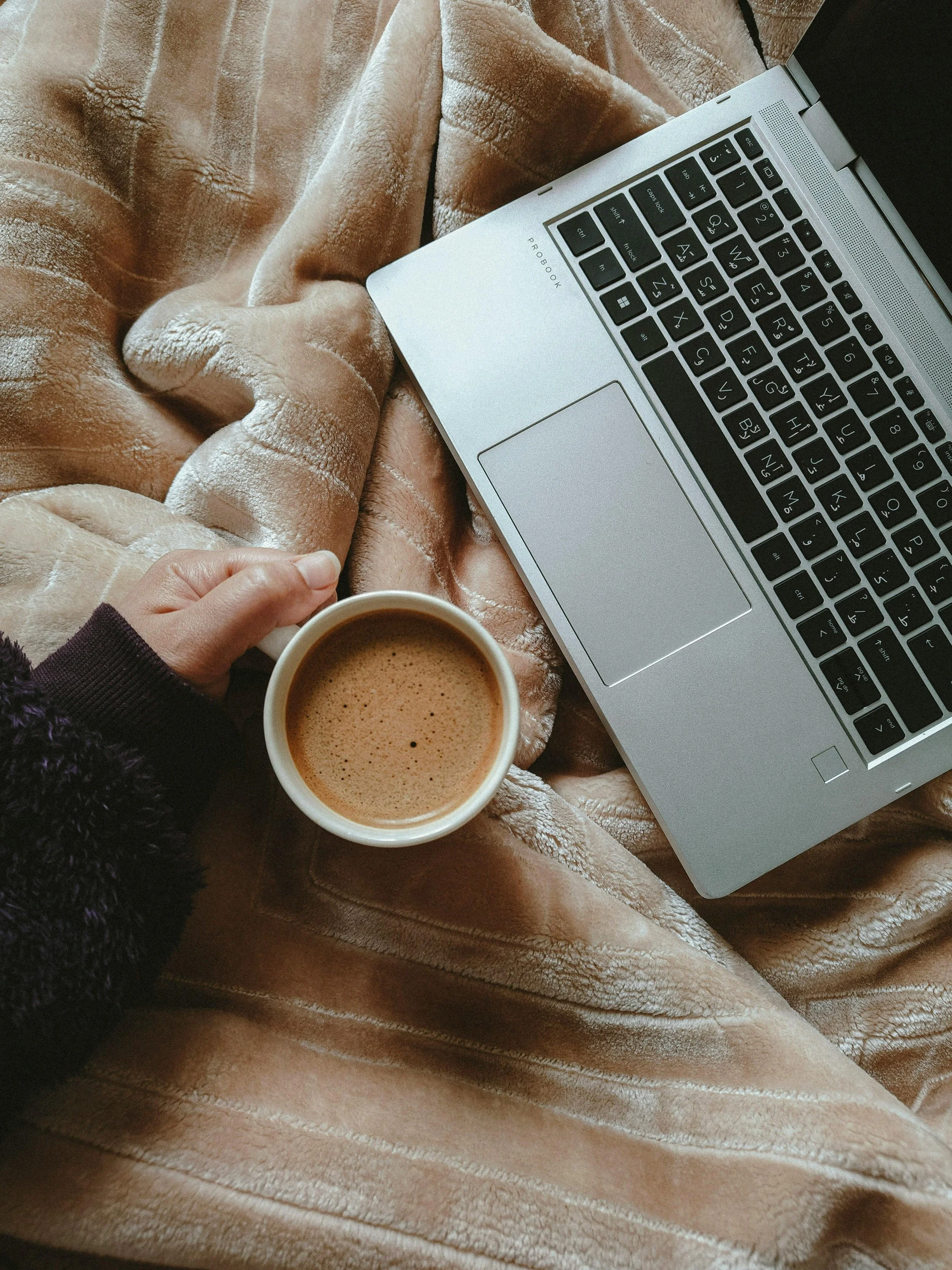 A person sitting on a cozy beige blanket, holding a cup of coffee, with a silver laptop placed nearby.