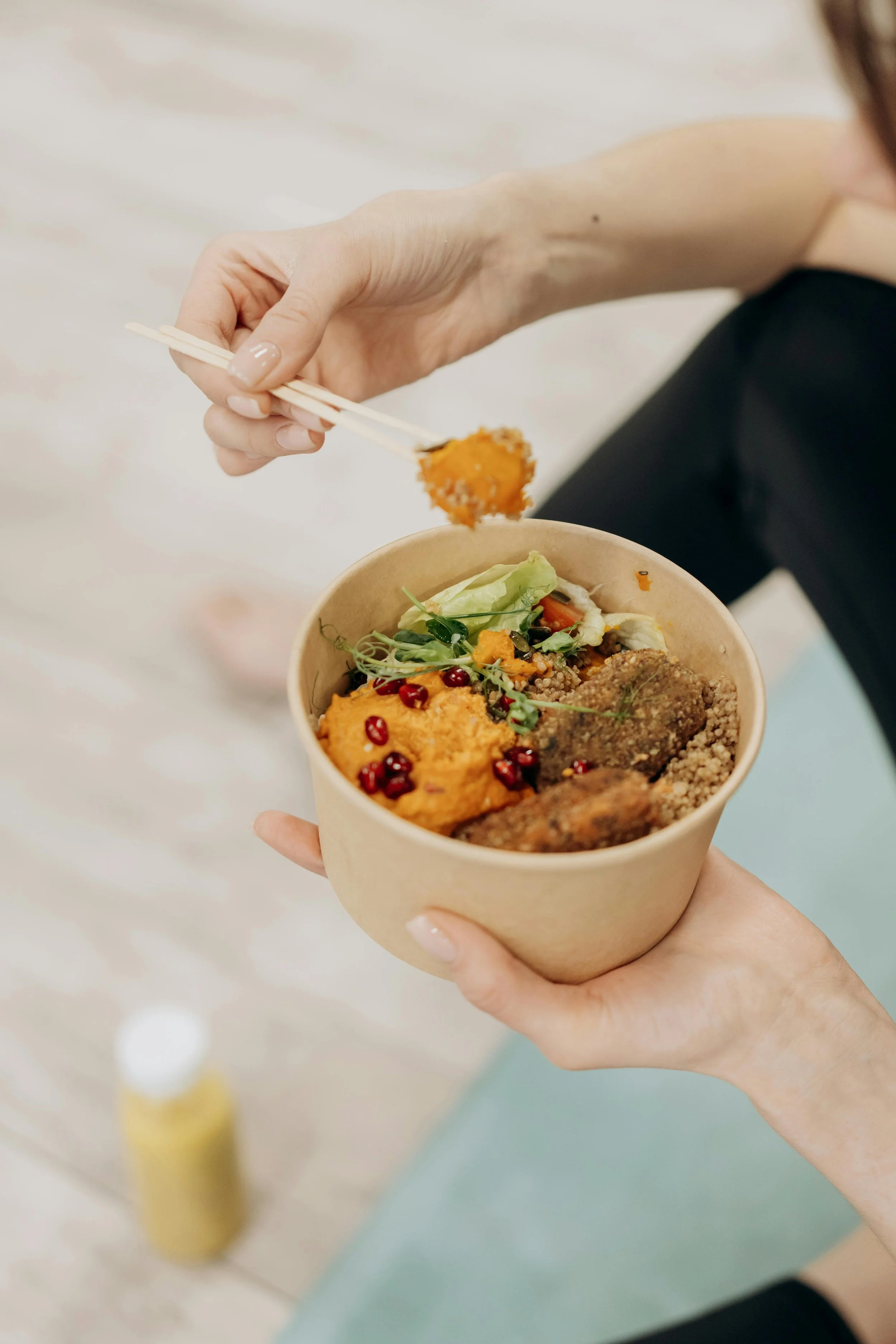 Person holding a cardboard bowl of salad and falafel, using chopsticks to pick up a piece of falafel.