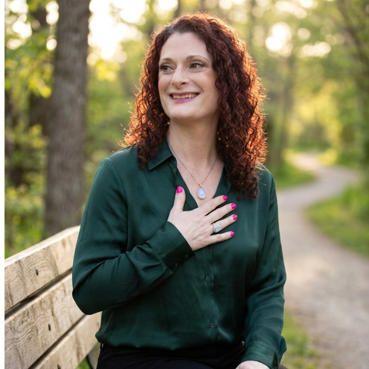 A woman with curly red hair sitting on a park bench, smiling with her hand on her chest, wearing a dark green blouse and jewelry, surrounded by trees and a pathway in the background.