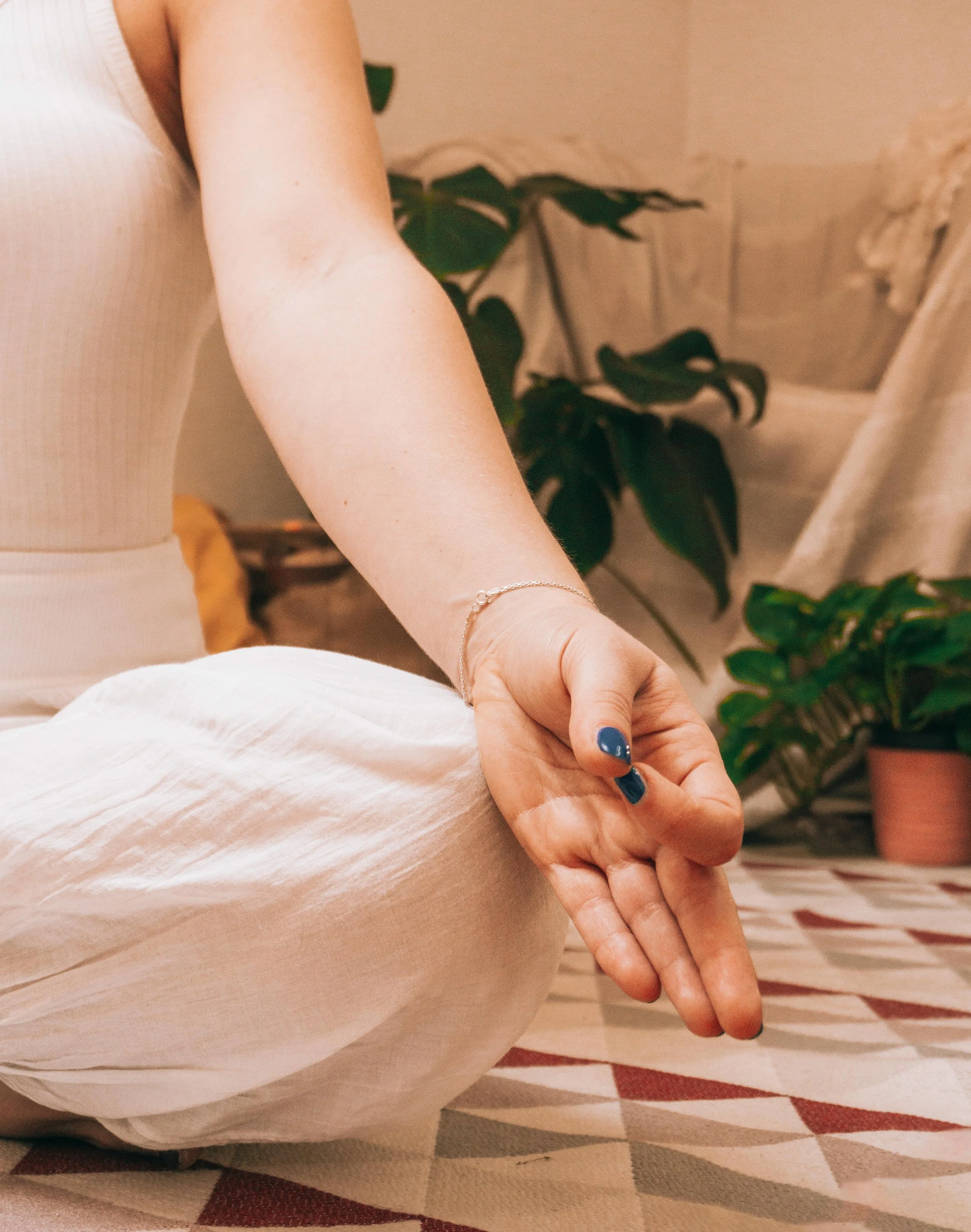 A person in a white outfit is sitting on a patterned rug with their hand resting on their knee, wearing a bracelet. In the background, there are green potted plants and a beige wall.