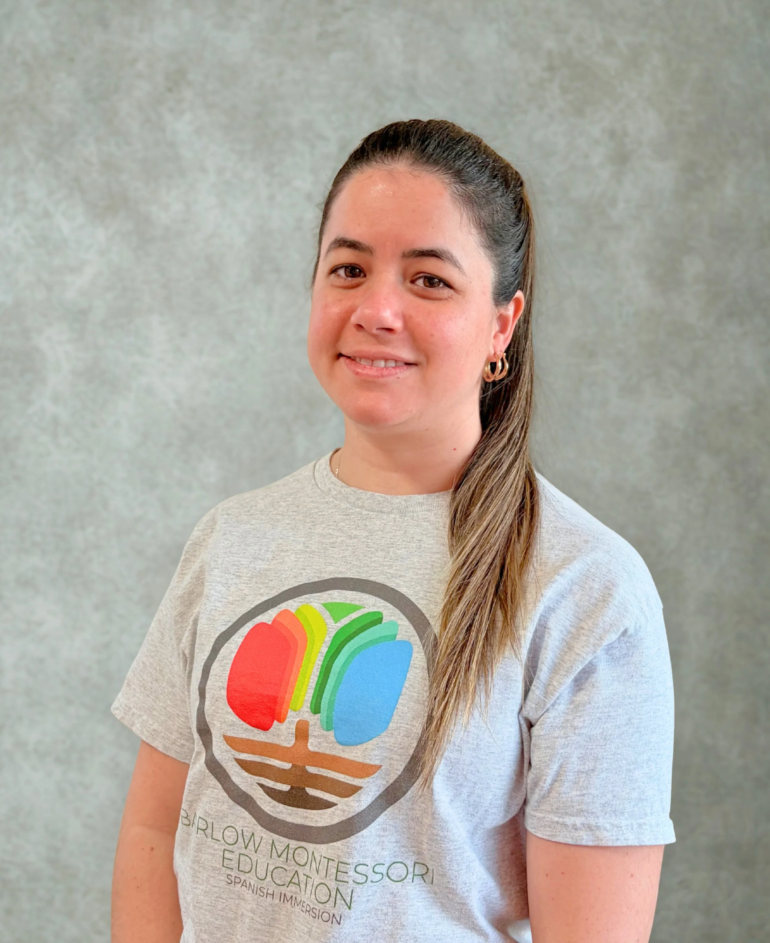 A smiling woman with long, wavy hair, wearing a white top, against a plain light gray background.
