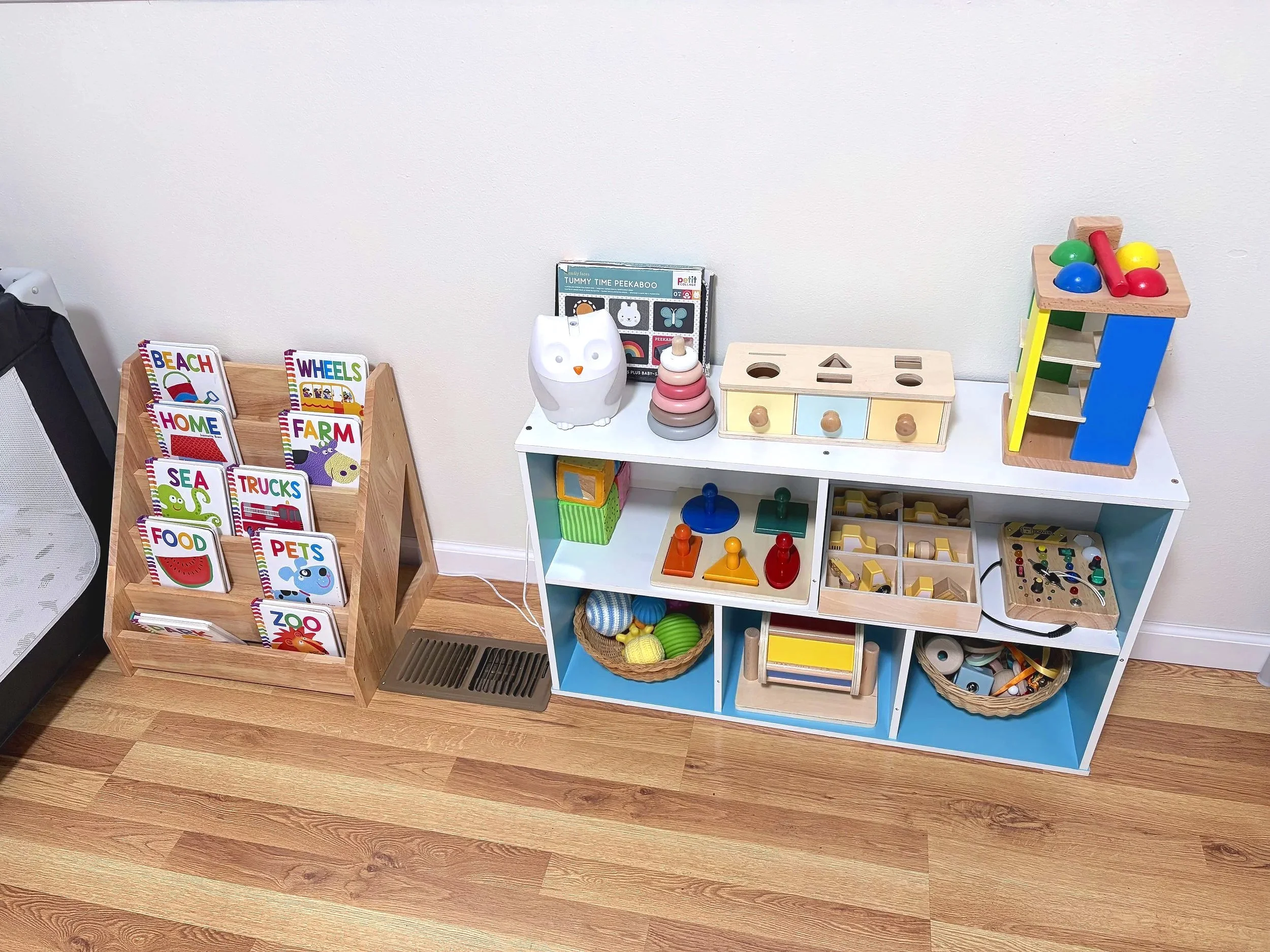 Children's toy display with wooden and plastic educational toys, books, and games on a white and blue shelf unit, and a wooden book holder on the floor with alphabet-themed books.