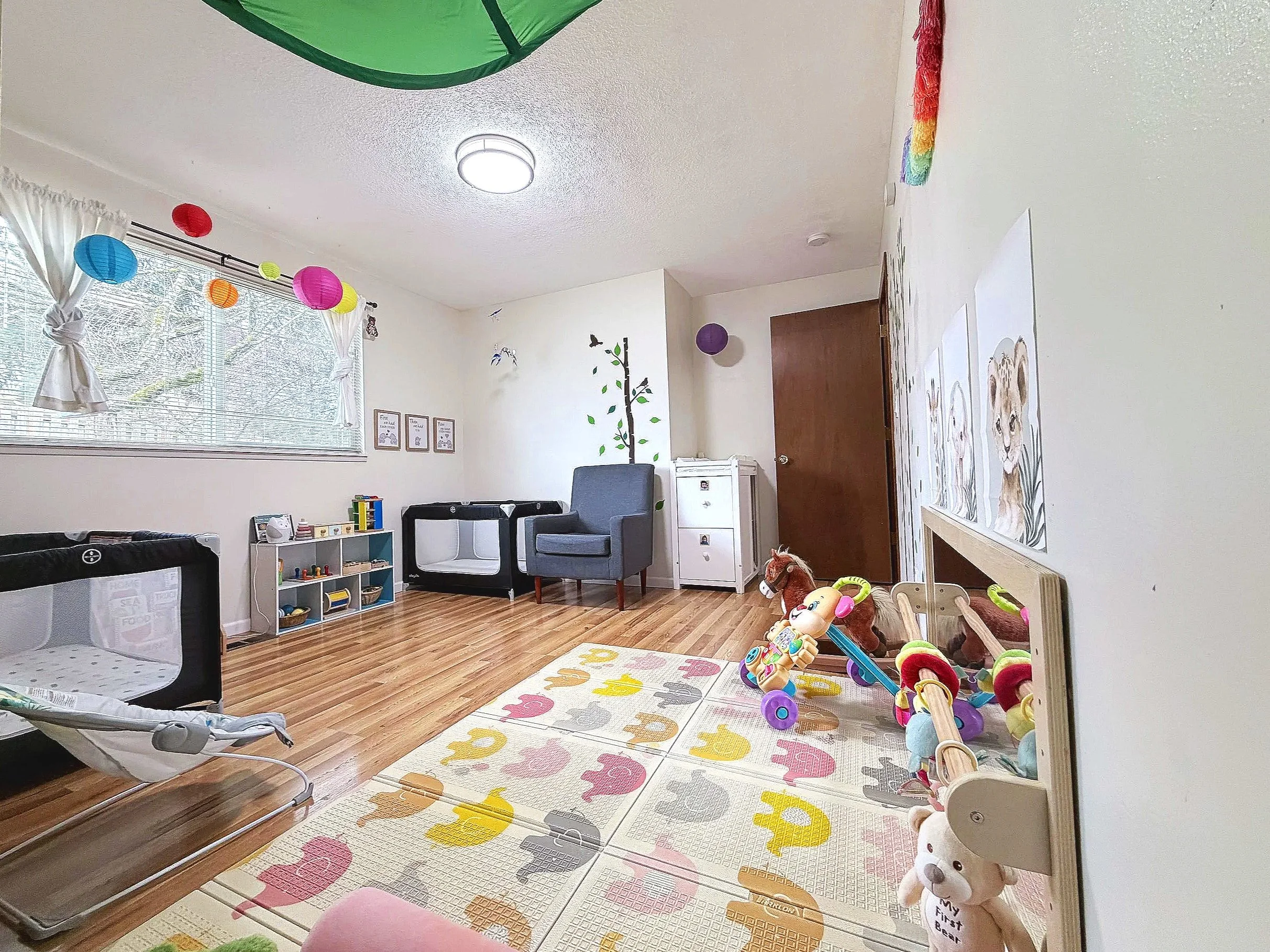 Children's playroom decorated with colorful paper lanterns hanging from the ceiling, a large window with white curtains, various toys including a stuffed dog and a wooden horse, a mirrored wall, and a multicolored elephant-themed rug.