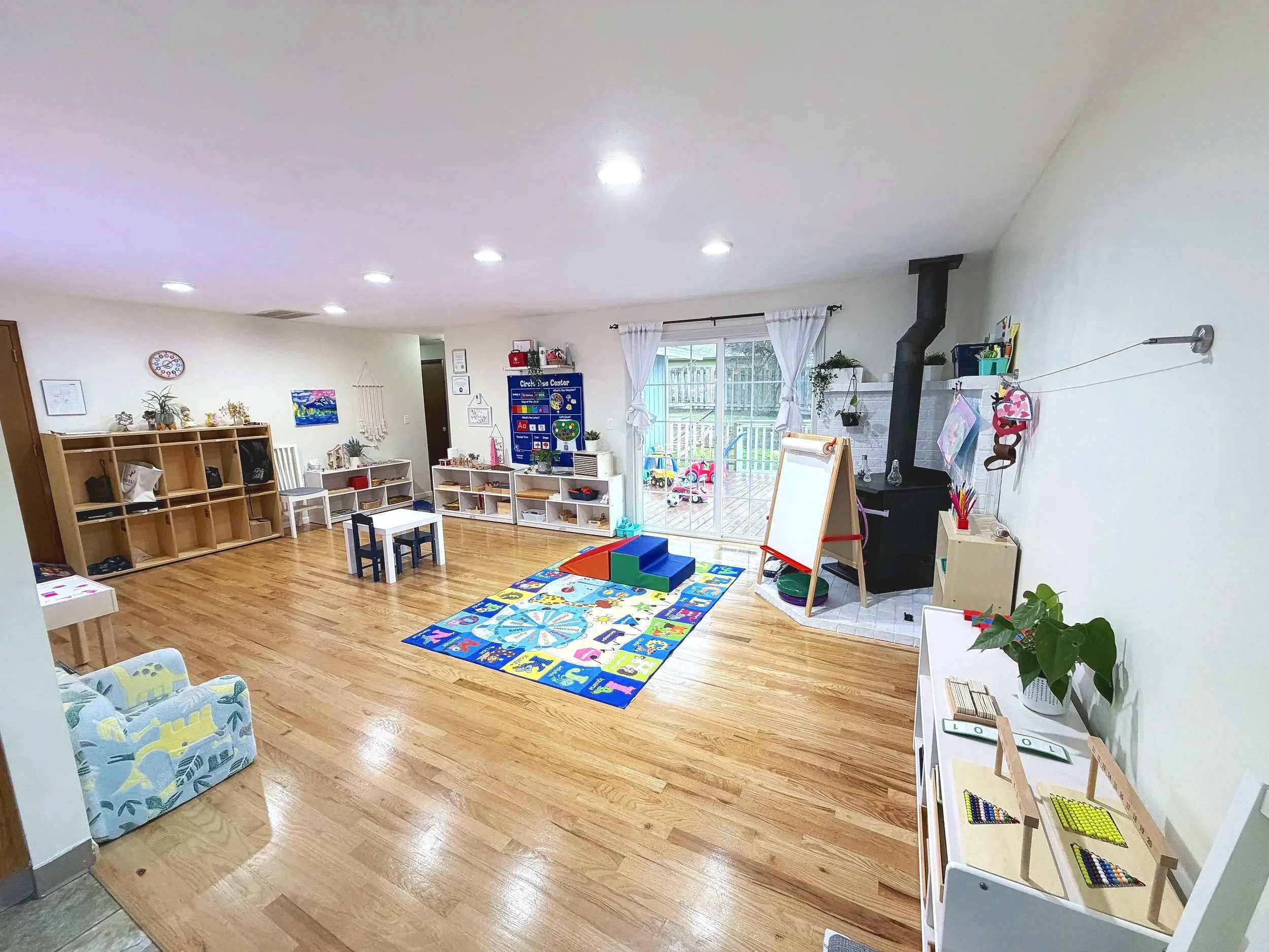 A spacious and well-lit children's playroom with wooden flooring, colorful toys, and educational materials, featuring a sliding glass door leading to an outdoor play area.