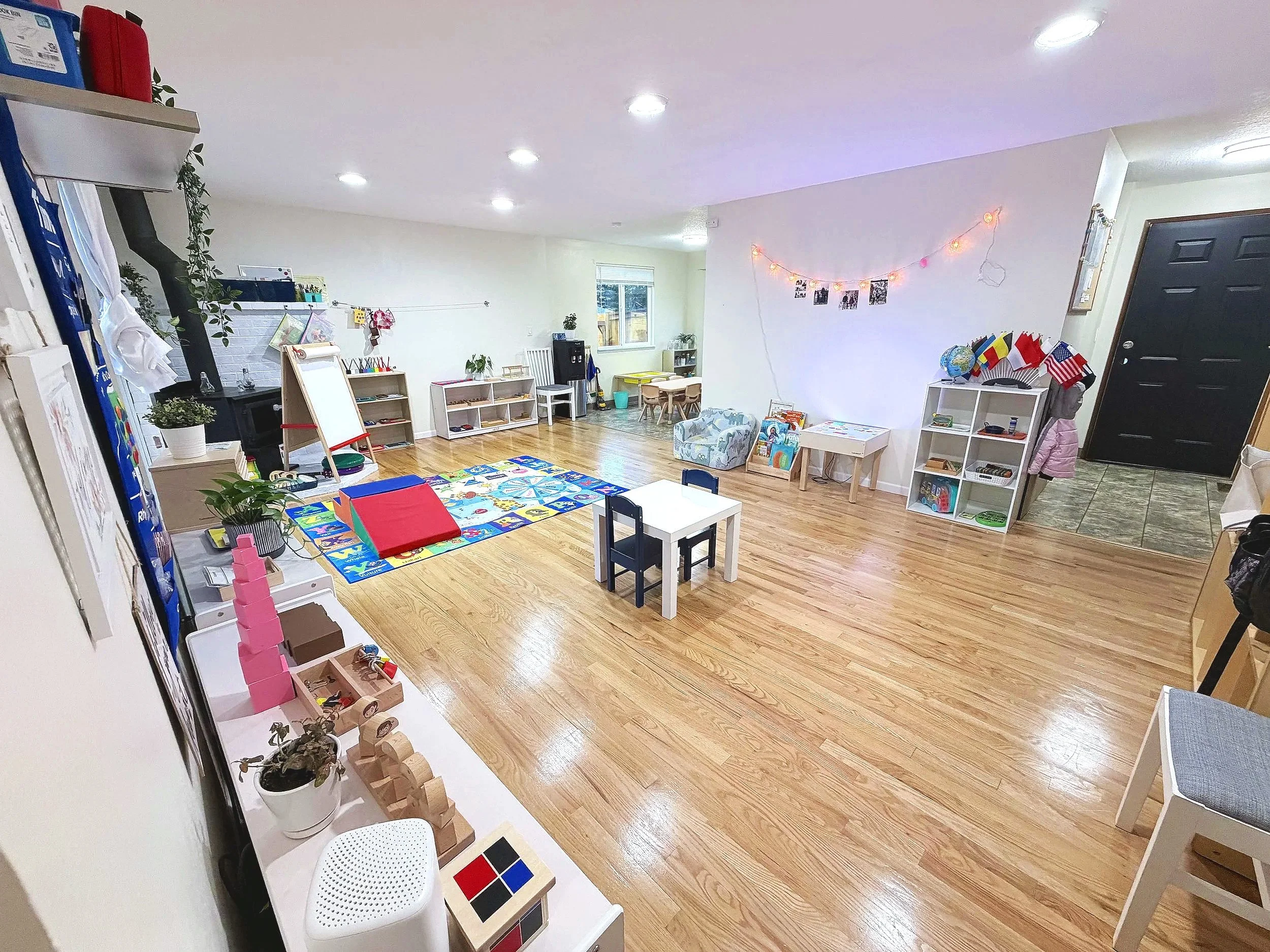 A spacious playroom with wooden floors, white walls, and various children's toys and furniture. There are small tables and chairs, a colorful play mat with toys, shelving units with books and toys, and decorations including string lights and flags. T