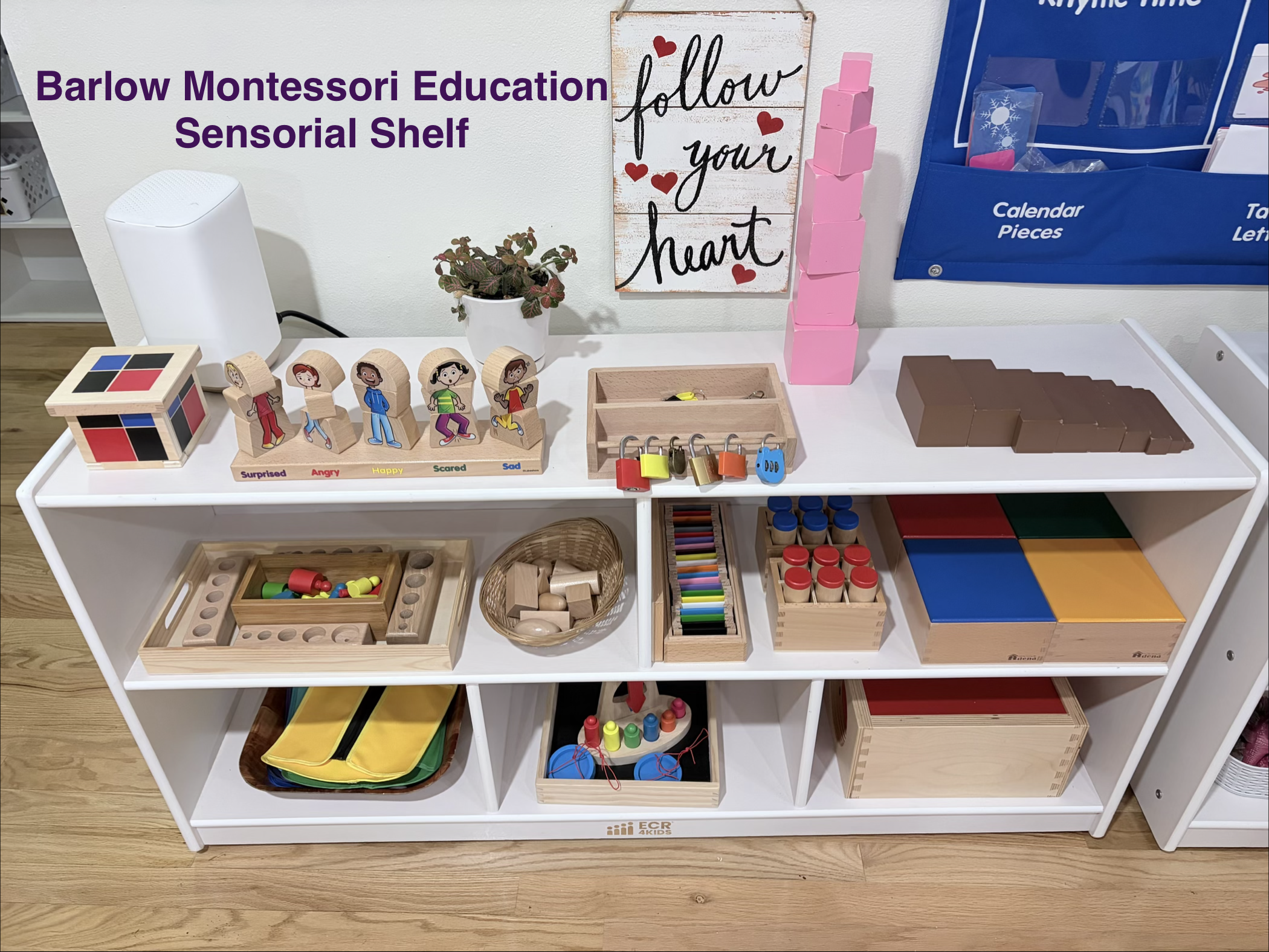 Montessori sensory shelf with educational toys including colored blocks, wooden figures with emotional labels, a lock set, paint squares, baskets with toys, and educational materials on white shelves in a classroom.