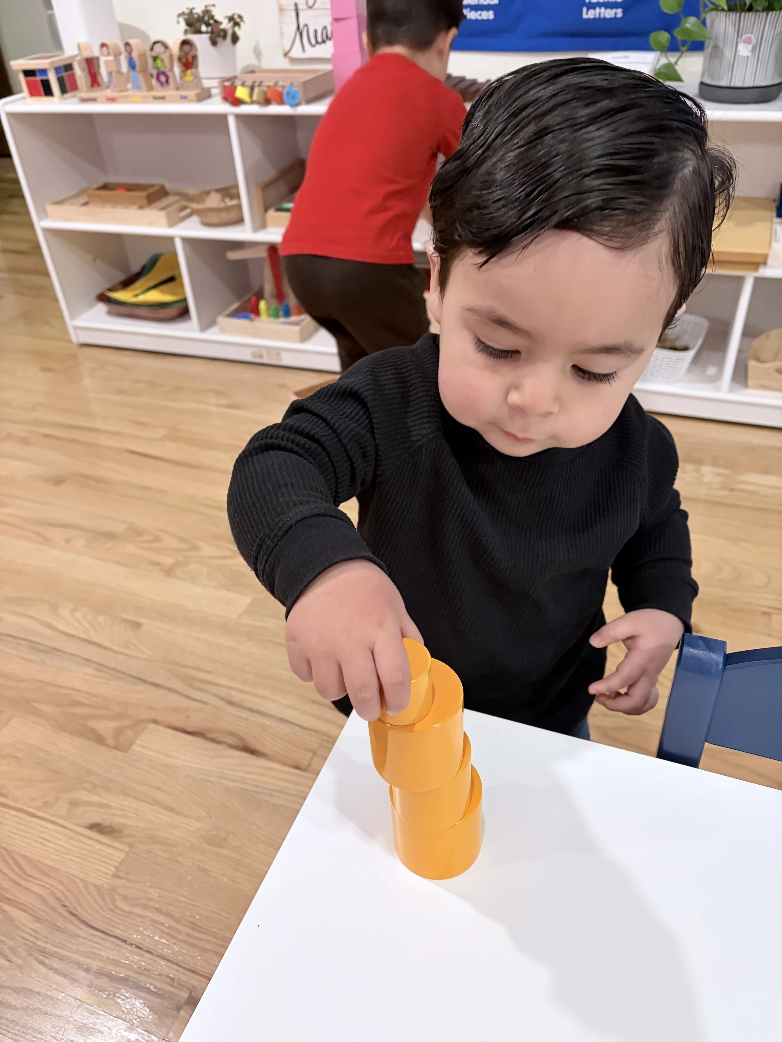 A young boy stacking orange plastic cups at a white table in a classroom or playroom setting.