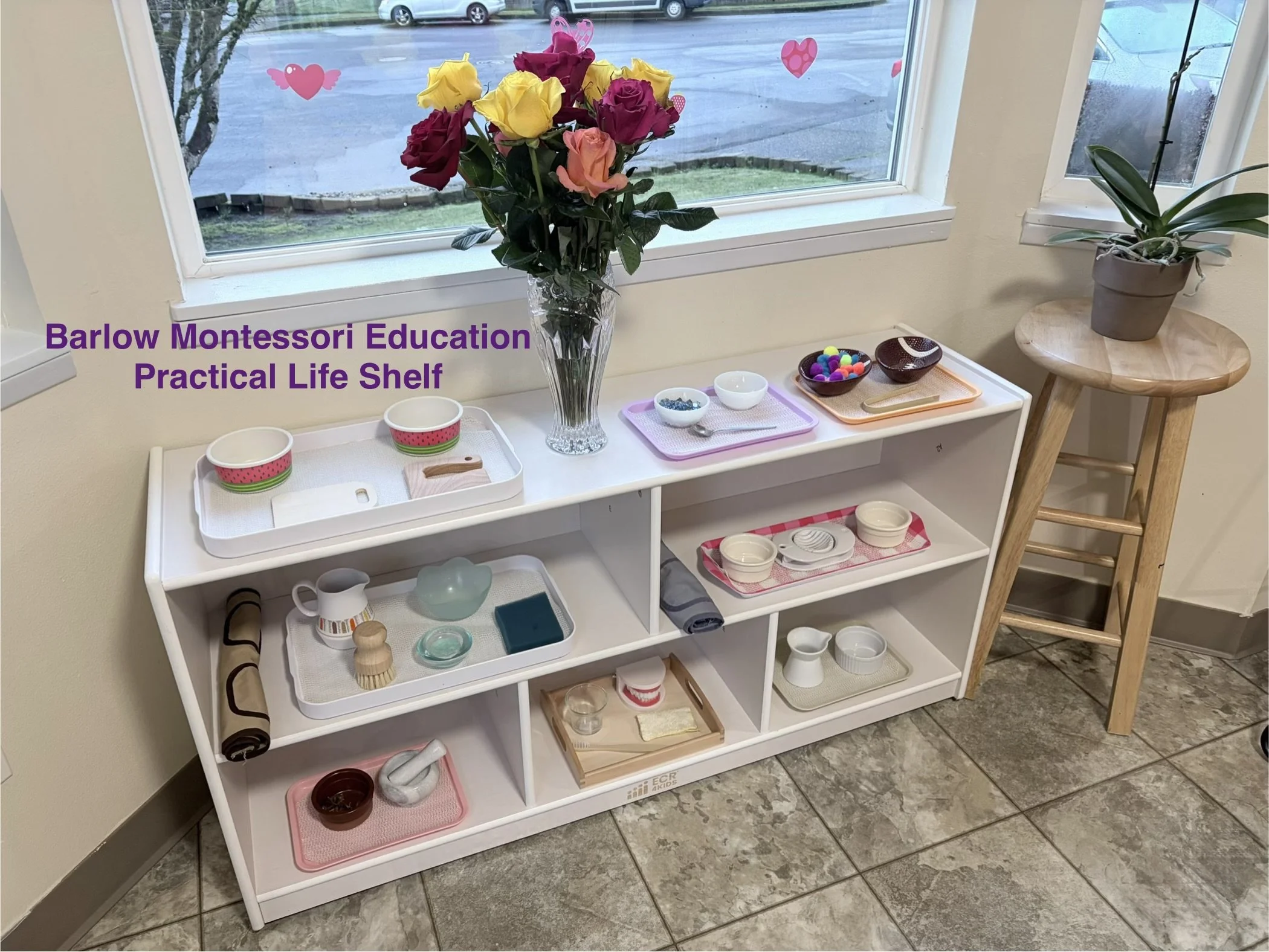 A white practical life shelf with various small items including bowls, a rolled mat, a pitcher, and wooden toys, with a vase of multicolored roses on top, positioned next to a window with pink heart decorations, and a potted plant on a wooden stool b