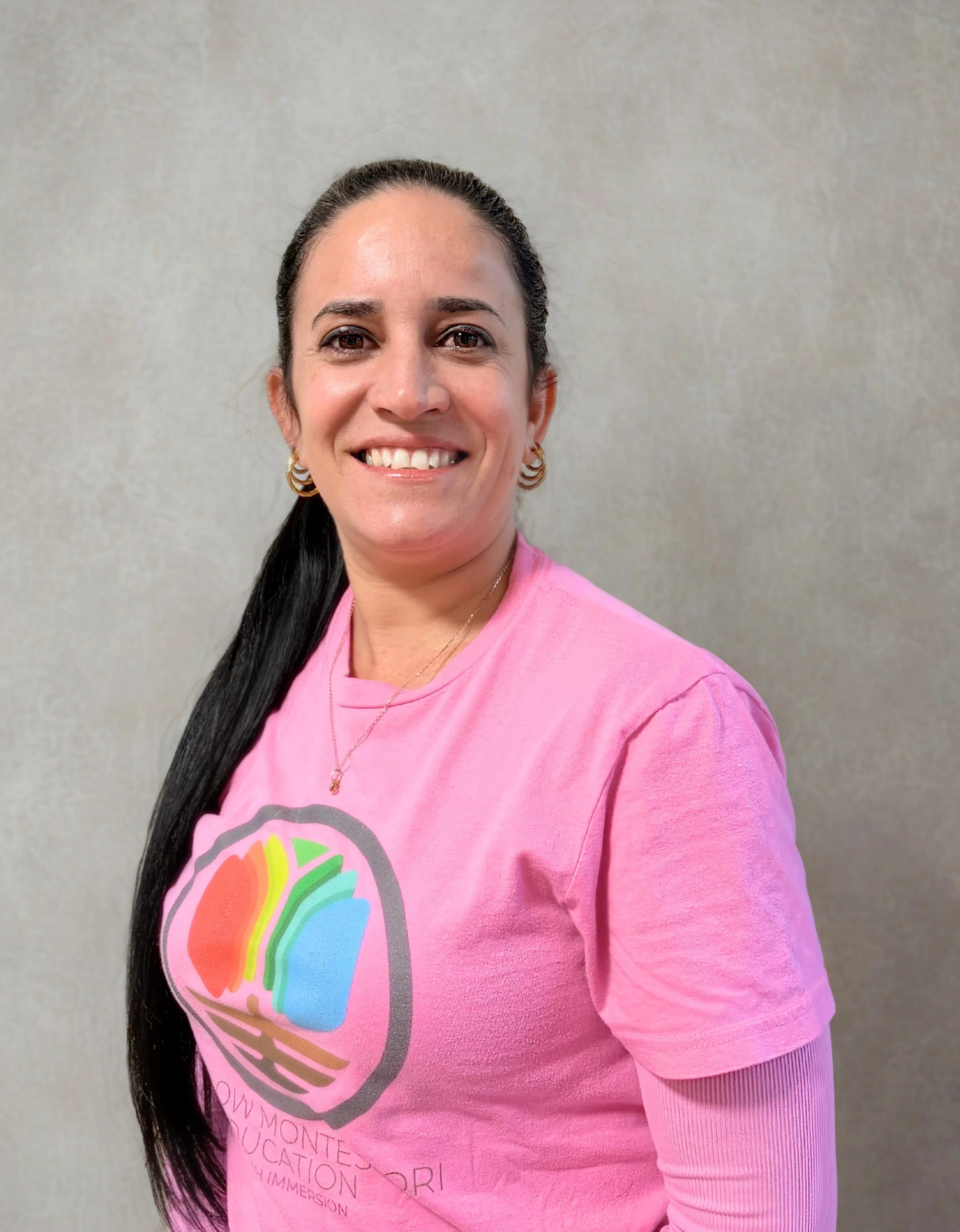 A woman with long dark hair tied back, smiling, wearing a black sleeveless top, gold earrings, and a gold necklace, standing against a plain light-colored wall.