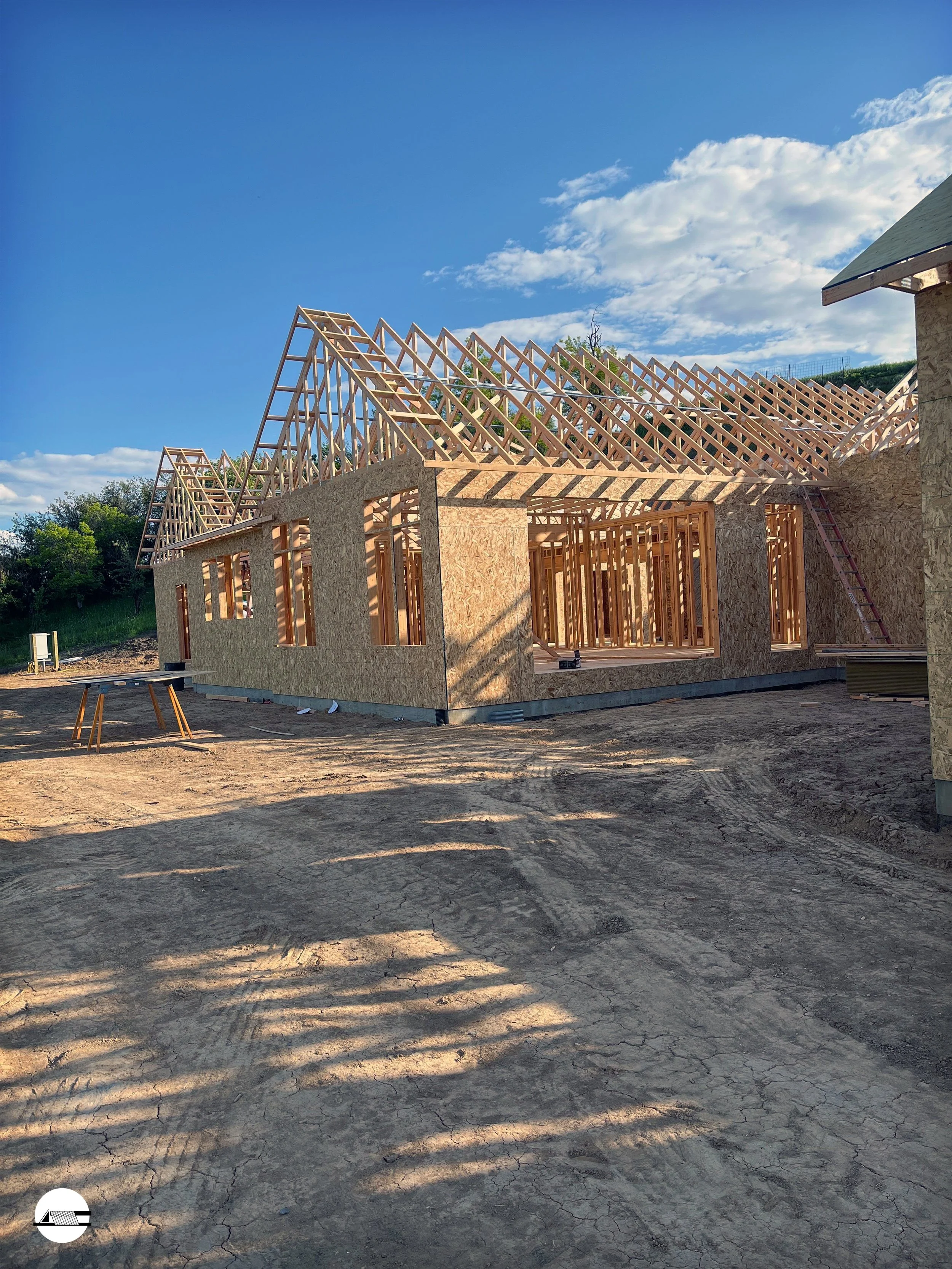 Wood framework for the roof of a house under construction, with walls made of plywood and open window spaces, on a dirt lot under a partly cloudy sky.
