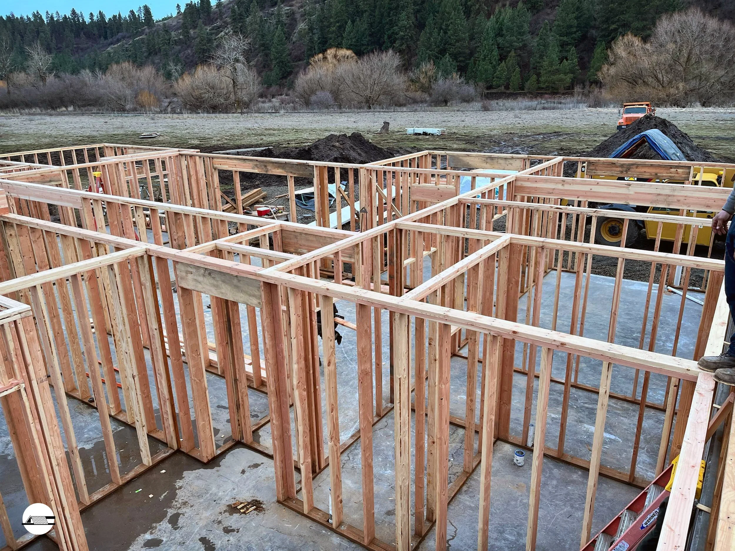 Construction site with wooden framing of a building in progress, set in a rural area with trees and hillside in the background.