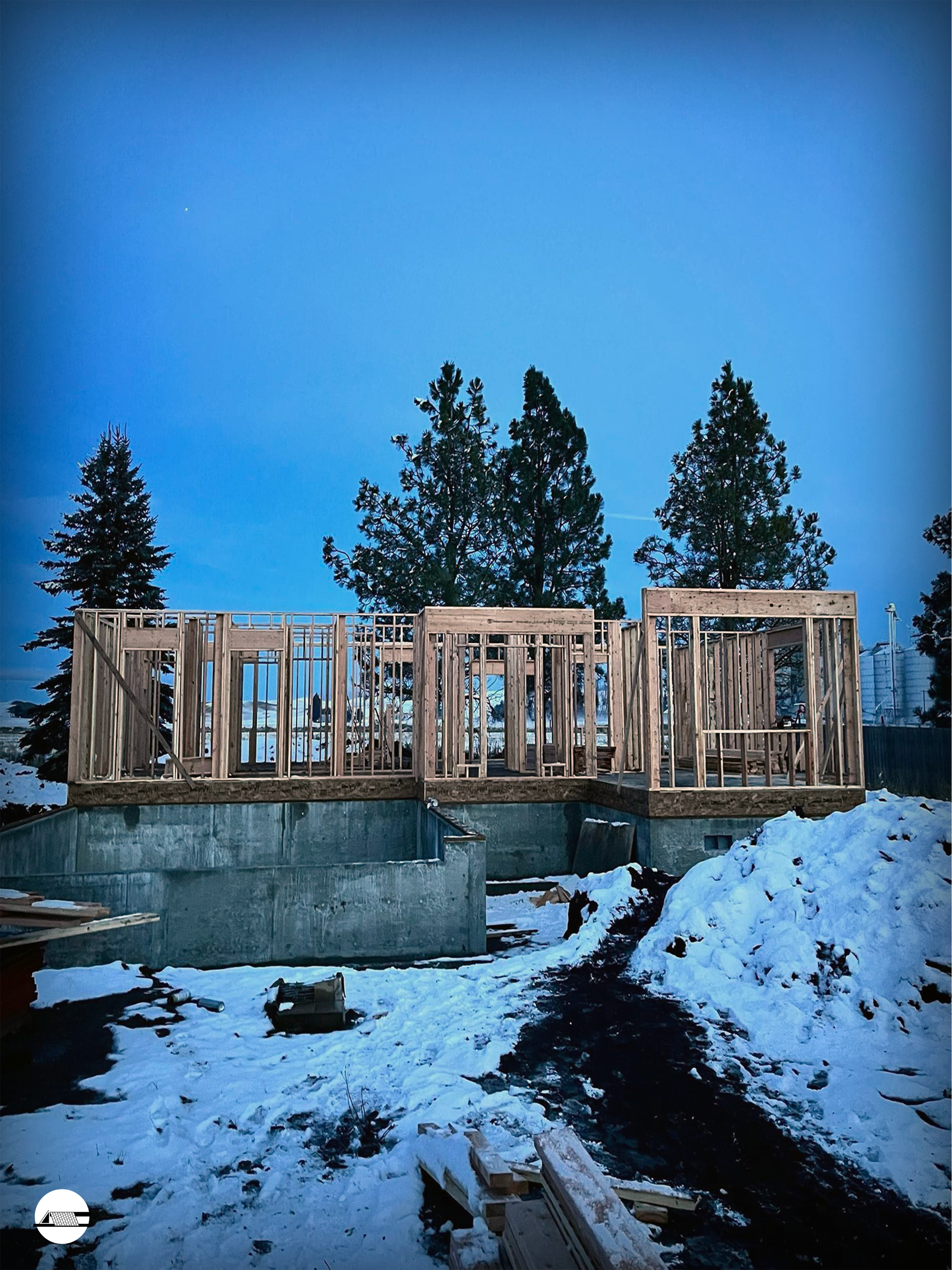 Construction site showing a wooden deck framework built on a concrete foundation, surrounded by snow with tall pine trees in the background during dusk or early evening.