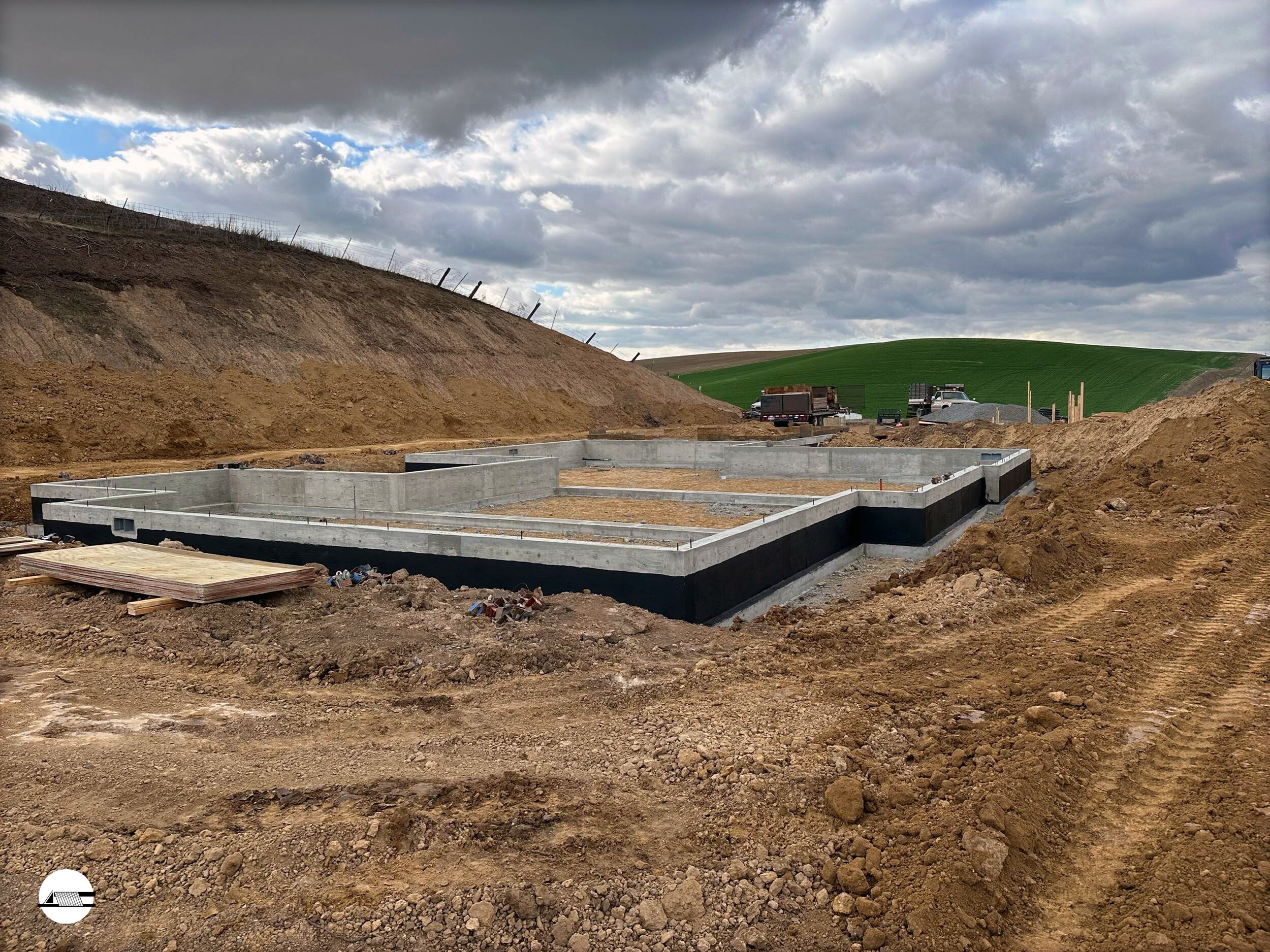 Construction site showing the foundation of a building with concrete walls, surrounded by dirt and construction equipment, under a cloudy sky with green fields in the background.