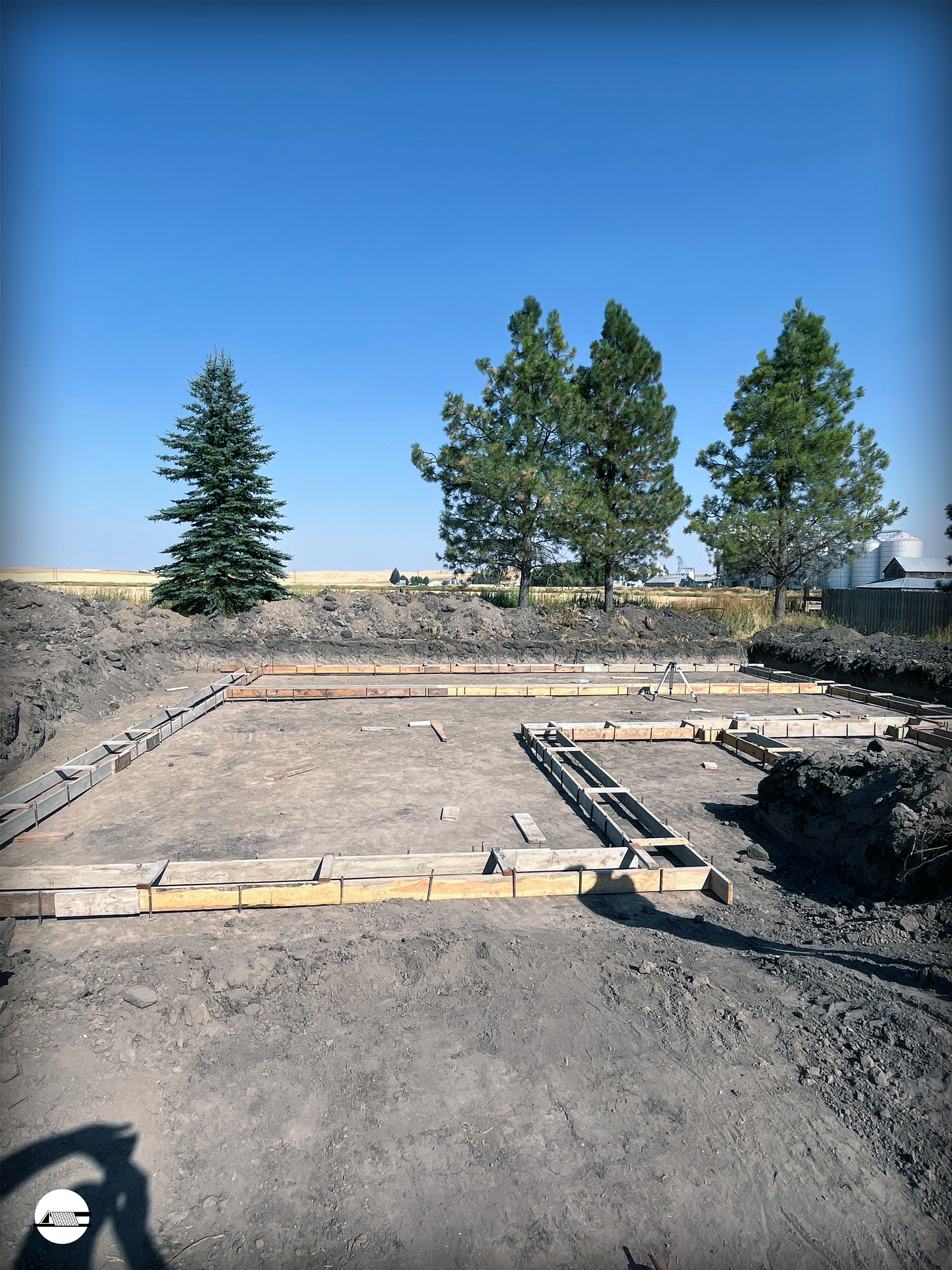 Construction site with foundation framework and three trees in the background under a clear blue sky.