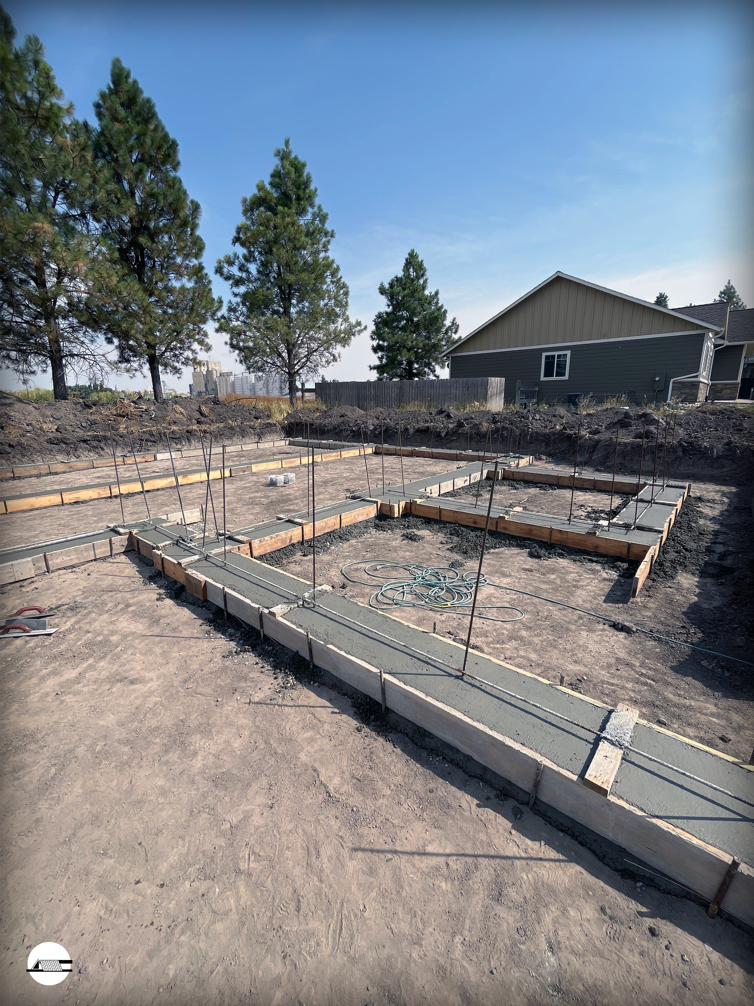 Construction site with concrete foundation and wooden formwork for a building, set against a background of green trees and a house with grey siding, under a clear blue sky.