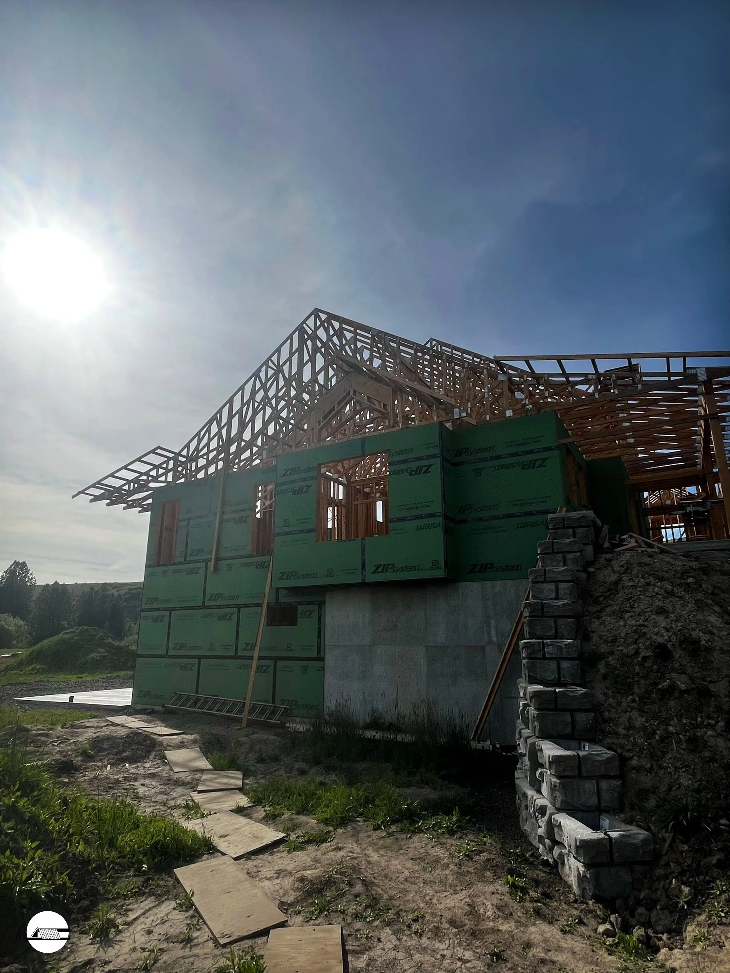 Under construction house with wooden framing, green exterior sheathing, and concrete foundation, with a stone staircase on the side. The sky is clear with the sun shining.