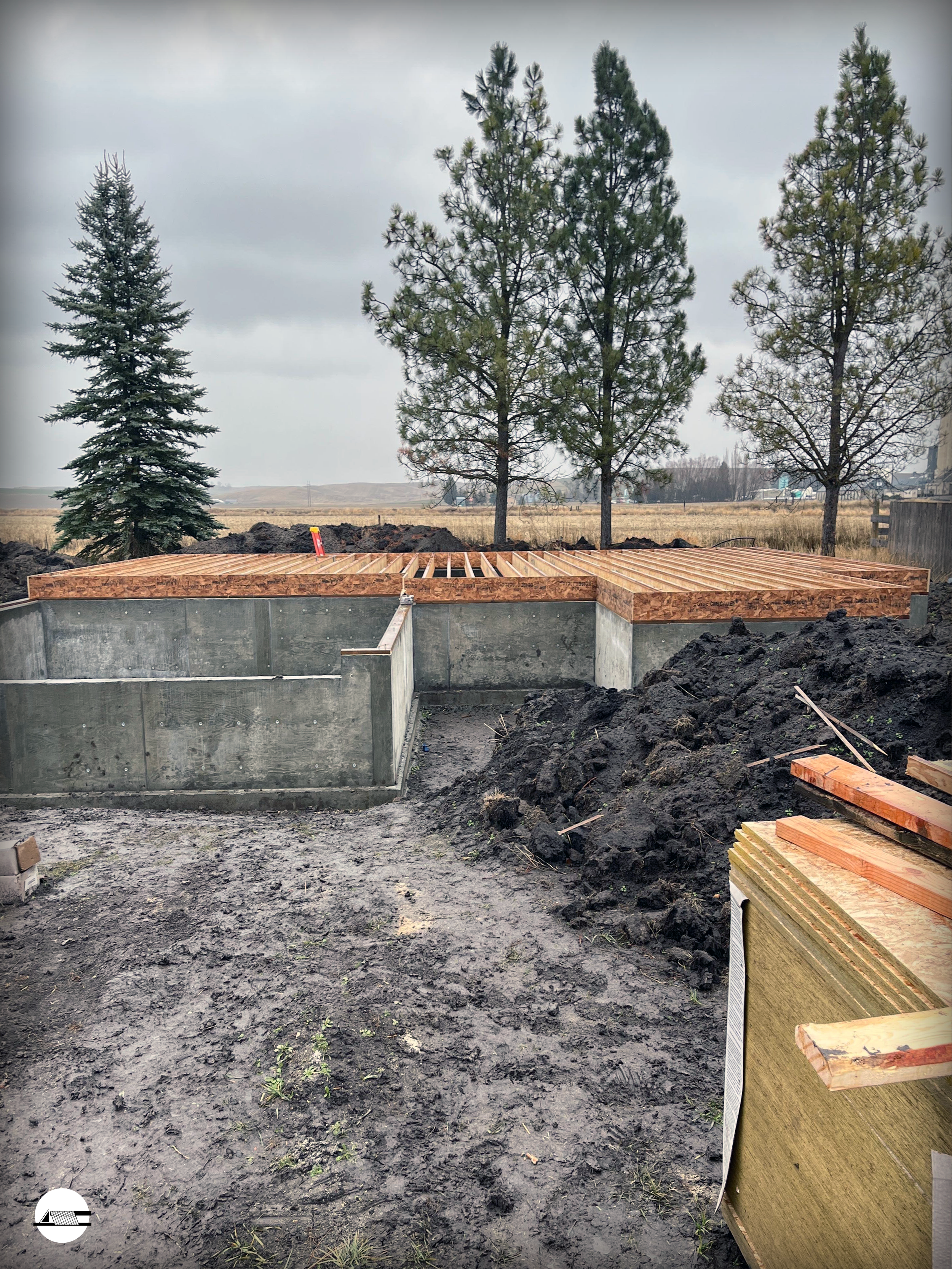 Construction site with a concrete foundation and wooden framing for a new building, surrounded by dirt and soil, with trees and cloudy sky in the background.