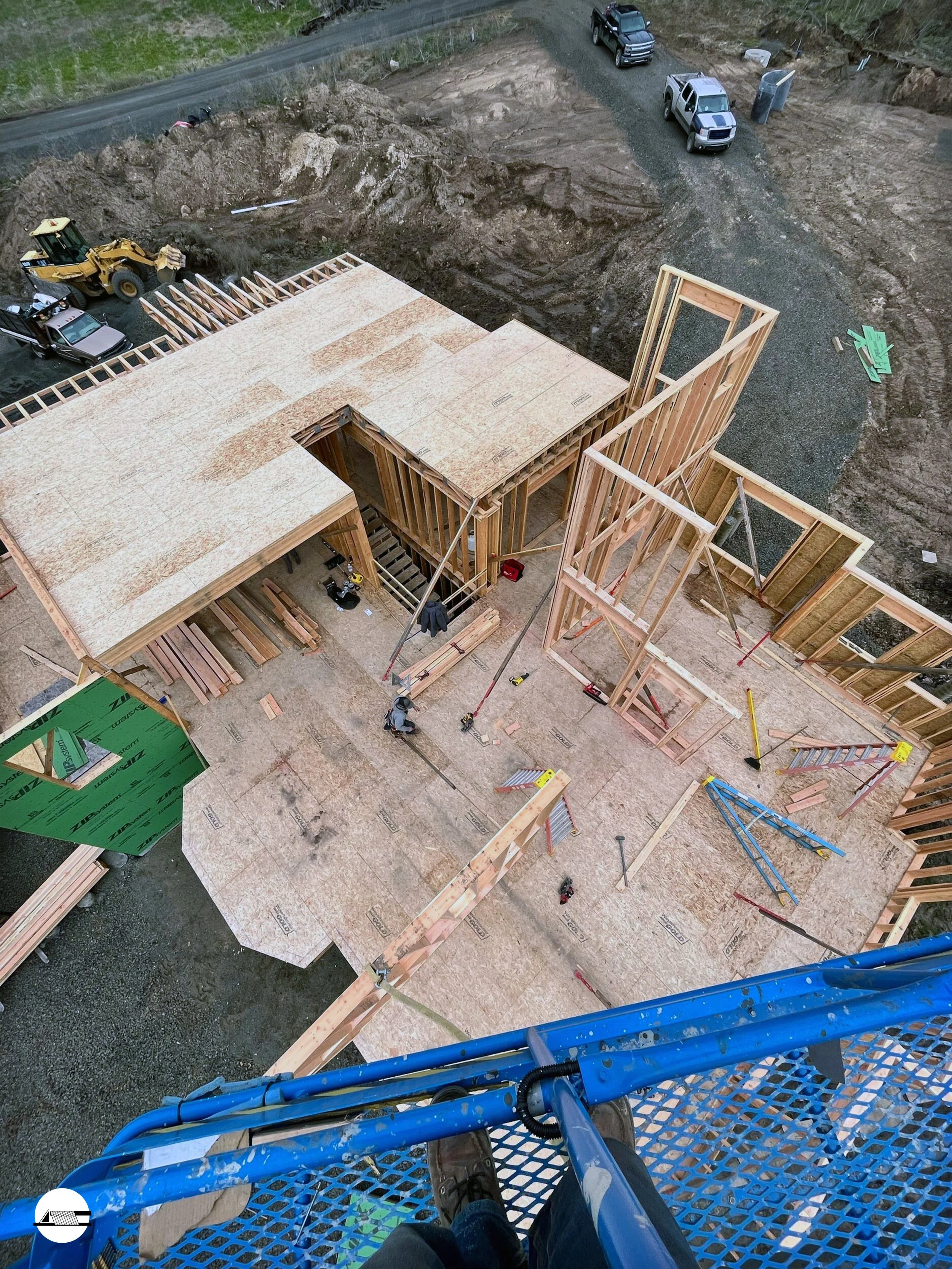 Aerial view of a house under construction with exposed wooden framework and flooring, surrounded by construction equipment and parked cars. The construction site is on a dirt lot with a gravel driveway and a road nearby.